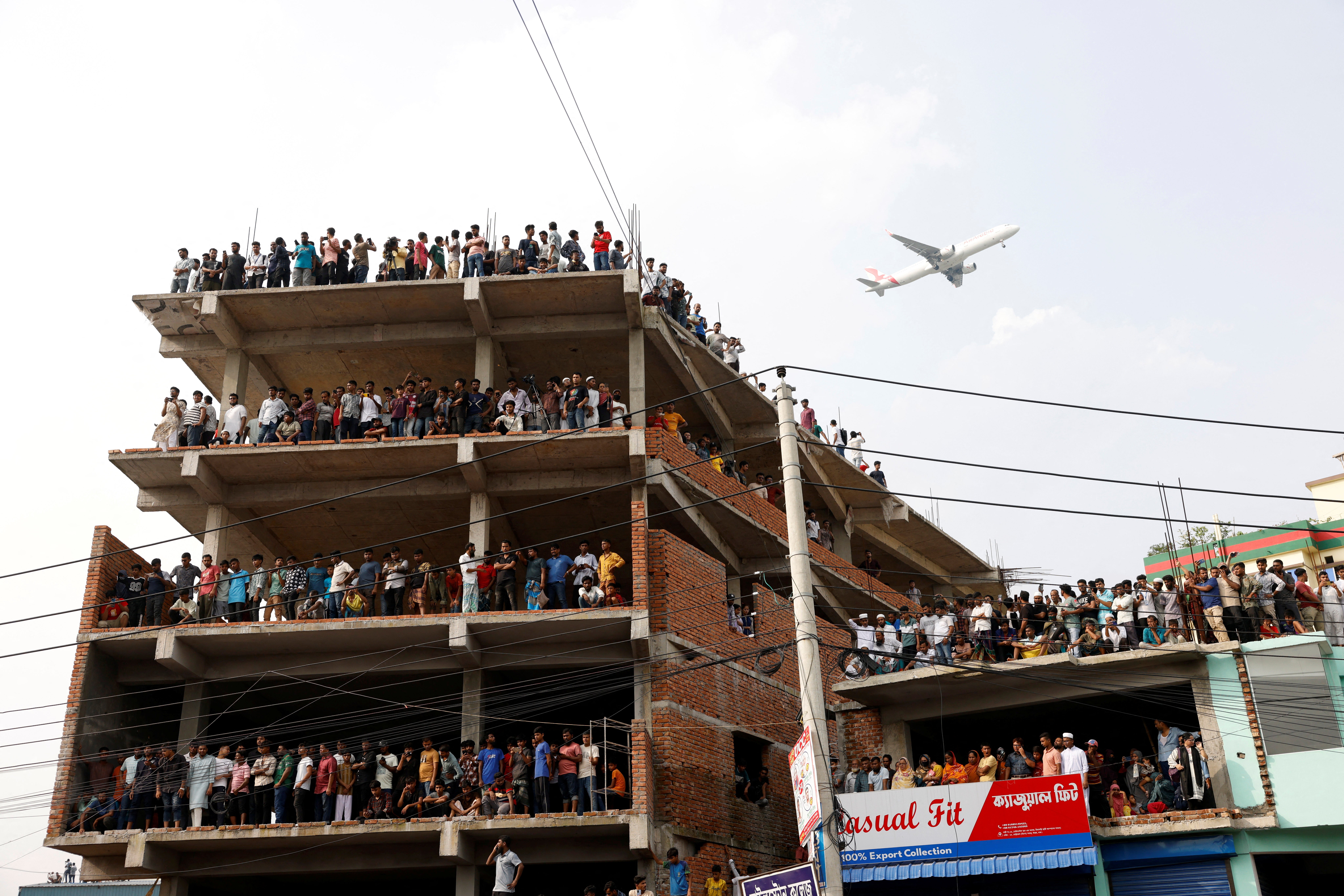 A passenger airplane flies as onlookers watch a rescue operation after an air force training aircraft crashed into Milestone College campus, in Dhaka