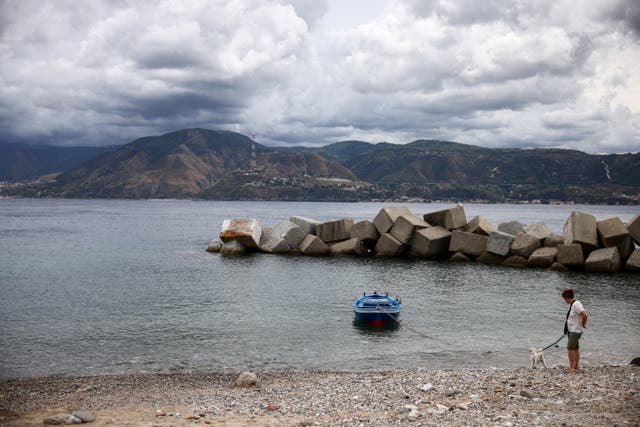 <p>A woman walks her dog along the Sicilian coast with the Calabrian shoreline visible across the Strait of Messina</p>