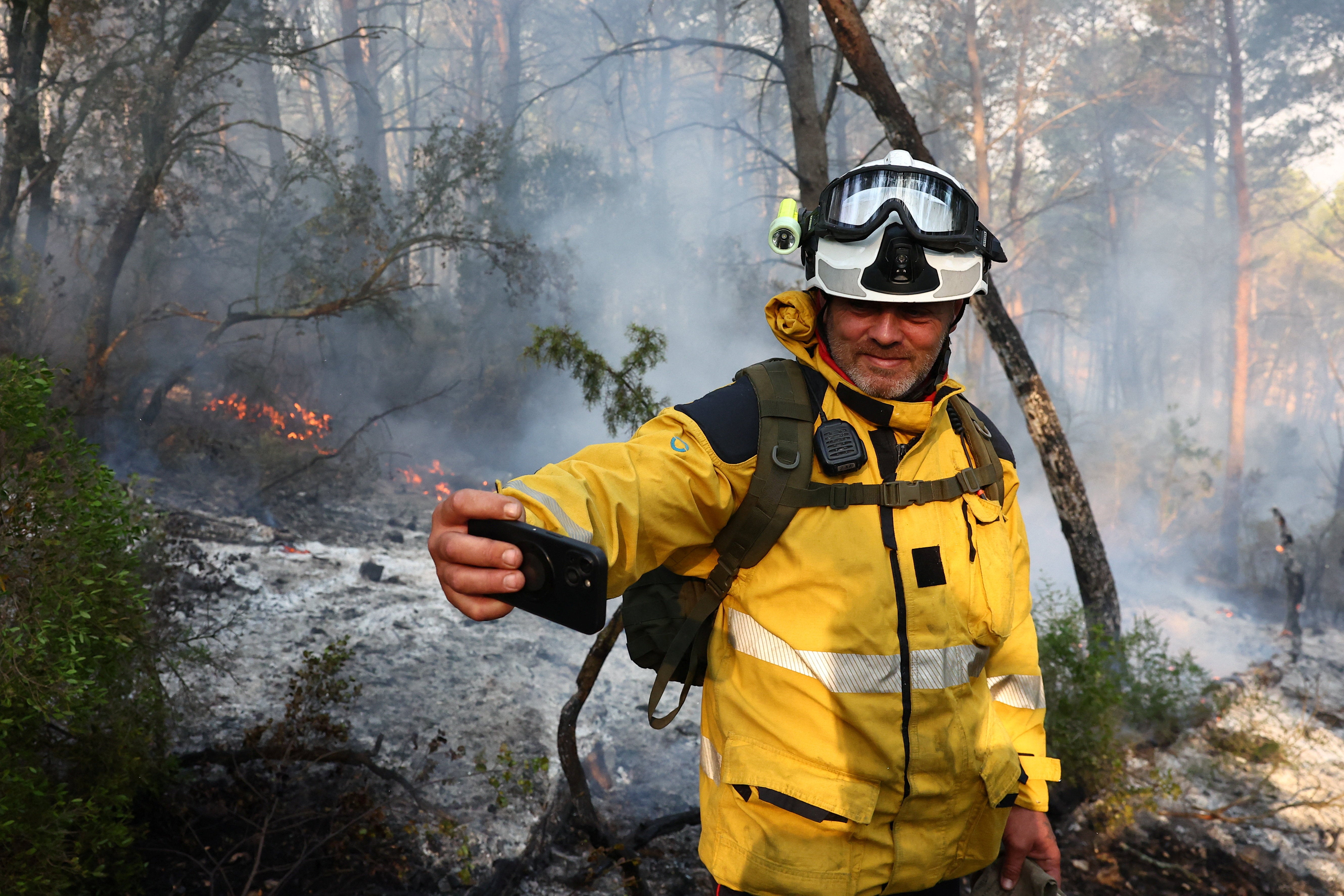A firefighter uses his phone as smoke rises from a wildfire burning in the background near Saint-Laurent-de-la-Cabrerisse, southern France, August 7, 2025. REUTERS/Abdul Saboor
