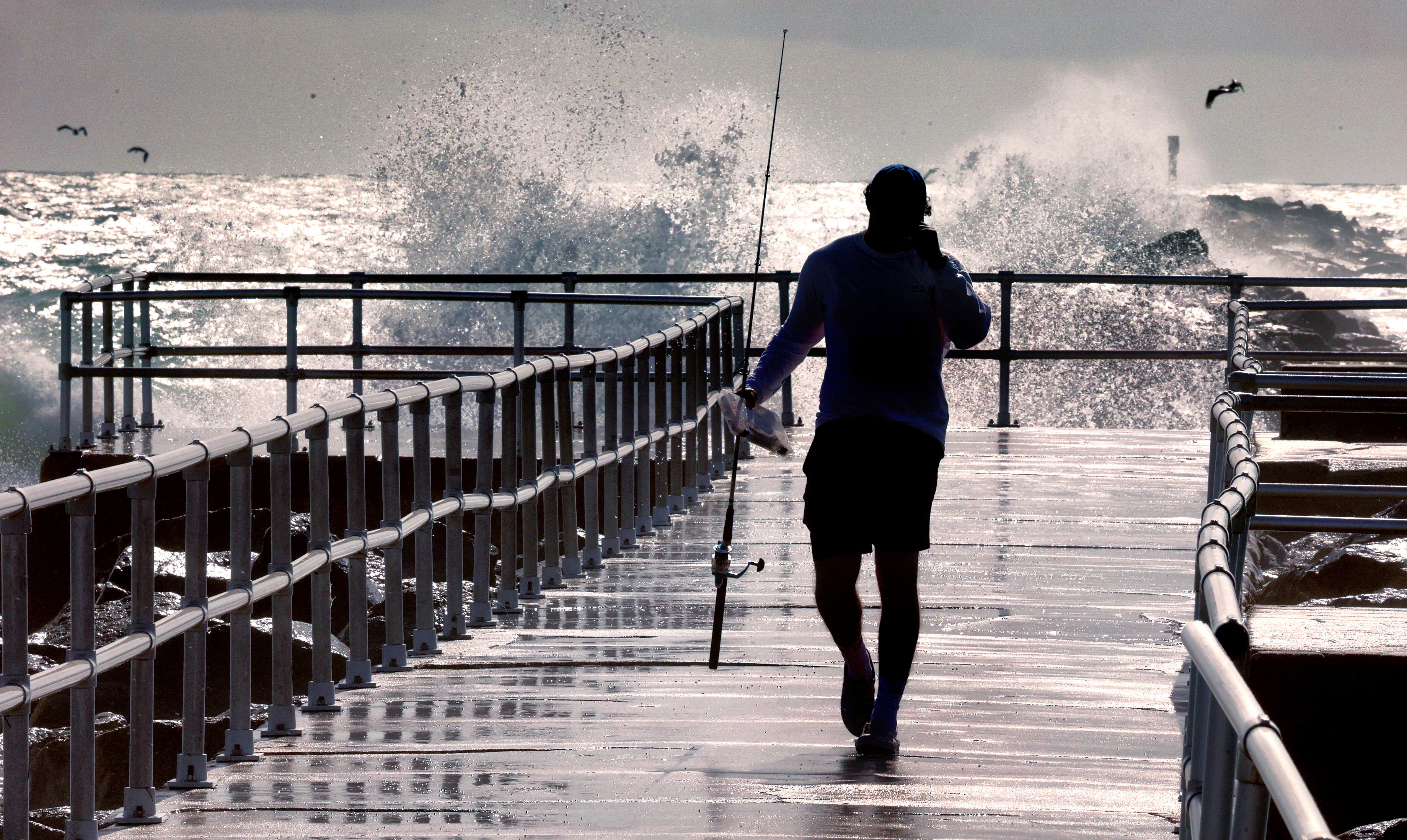 A fisherman walks on a pier as large waves generated by Hurricane Erin crash into the jetty at Lighthouse Point Park, in Ponce Inlet, Florida
