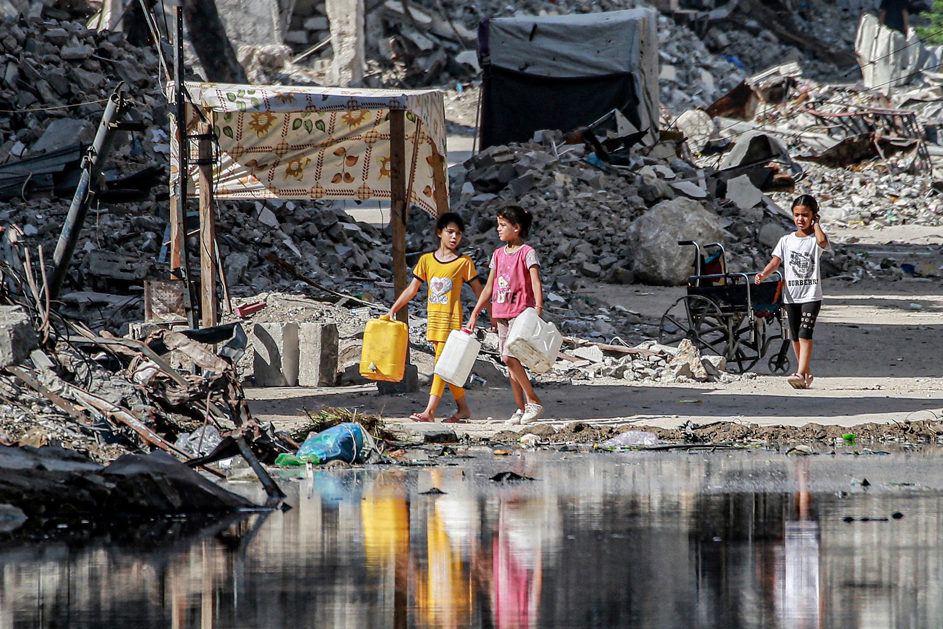 Children walk past destroyed buildings and a puddle of sewage water erupting from collapsed underground pipes in Khan Younis