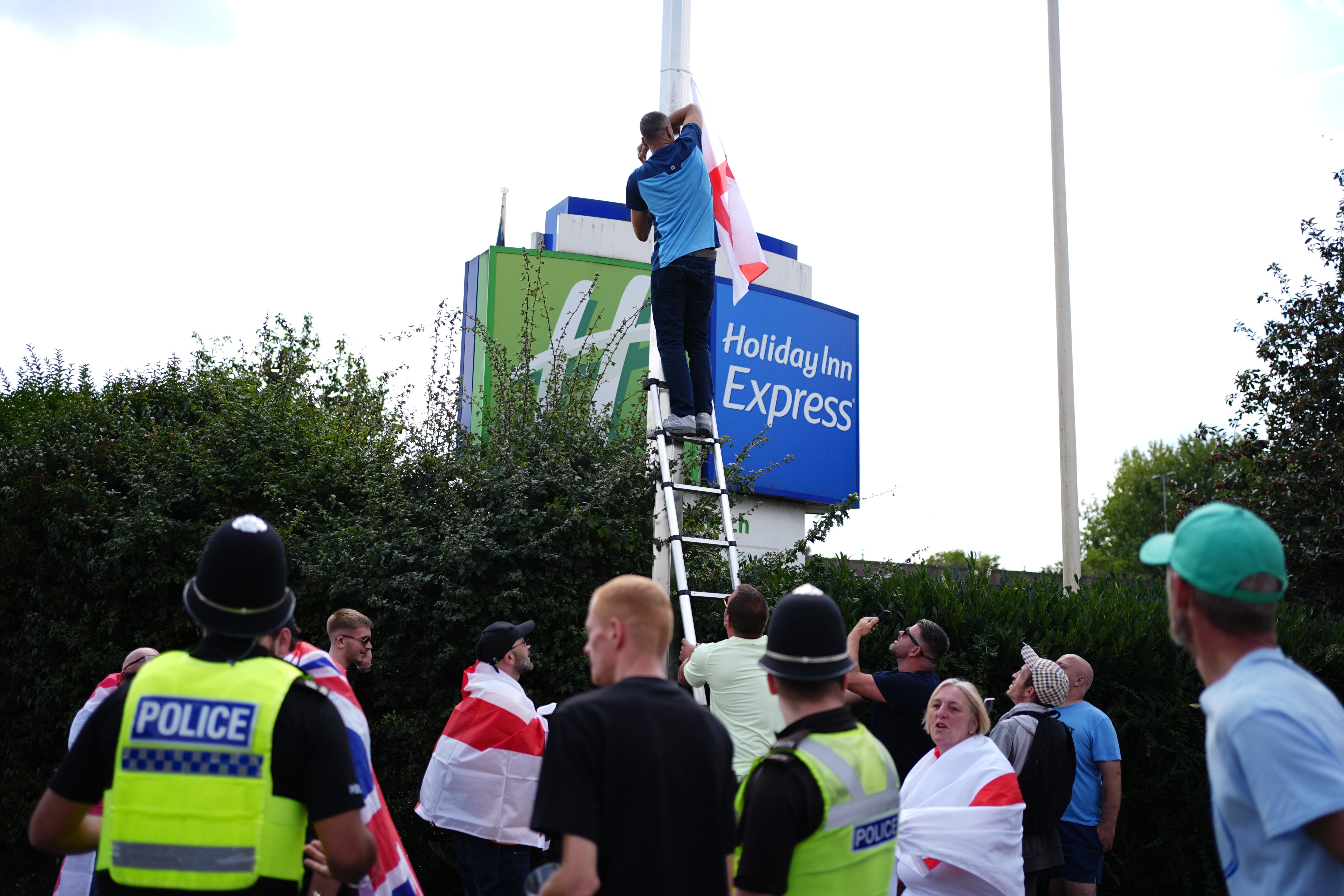 A man puts up a St George’s Cross flag on a lamp post as people demonstrate under the Abolish Asylum System slogan outside the Castle Bromwich Holiday Inn in Birmingham (Jacob King/PA)