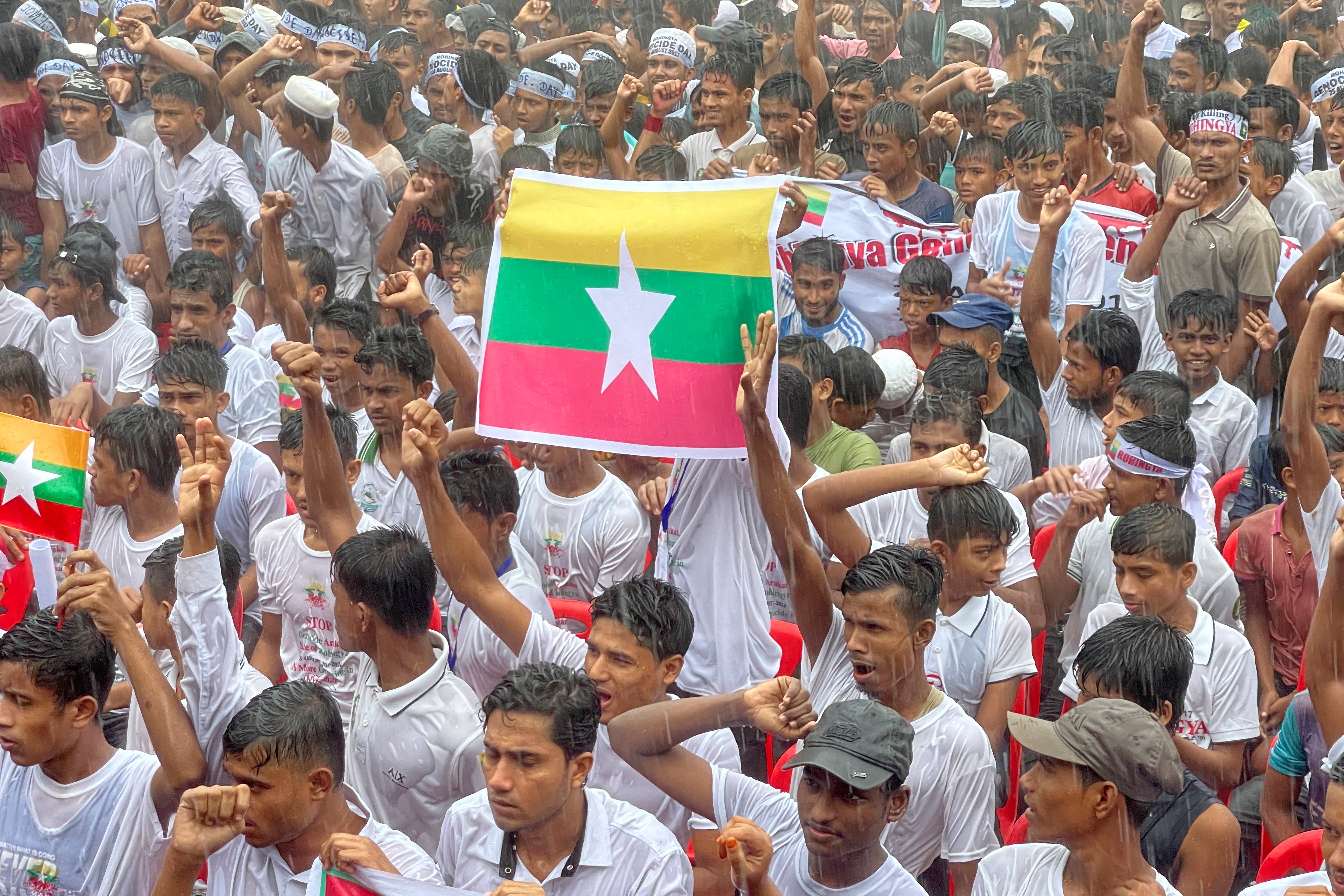 File: Rohingya refugees gather in the rain, with a flag of Myanmar, center, to demand safe return to Myanmar's Rakhine state as they mark the seventh anniversary of their mass exodus at their refugee camp at Kutupalong in Cox's Bazar district, Bangladesh