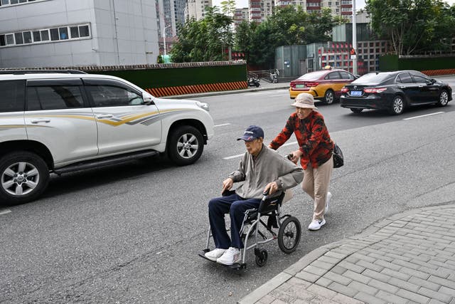<p>A woman pushes an elderly man in a wheelchair along a street in Beijing</p>