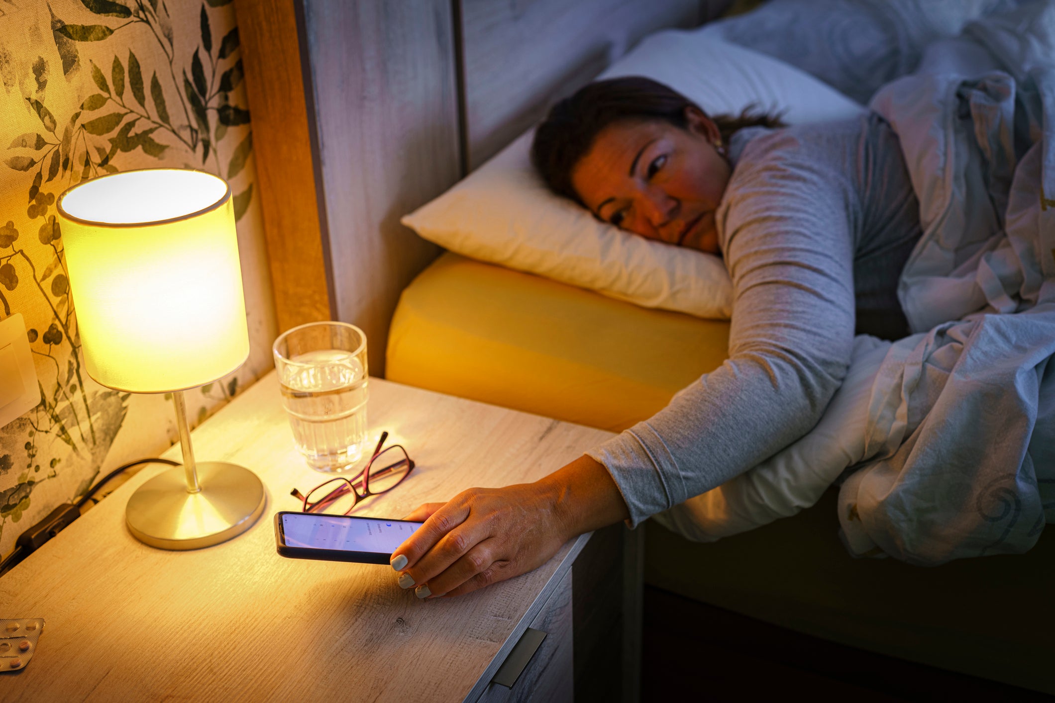 <p>Close up of mature woman laying on bed reaching her mobile phone on the night table early in the morning. Selective focus on hand and clock. </p>