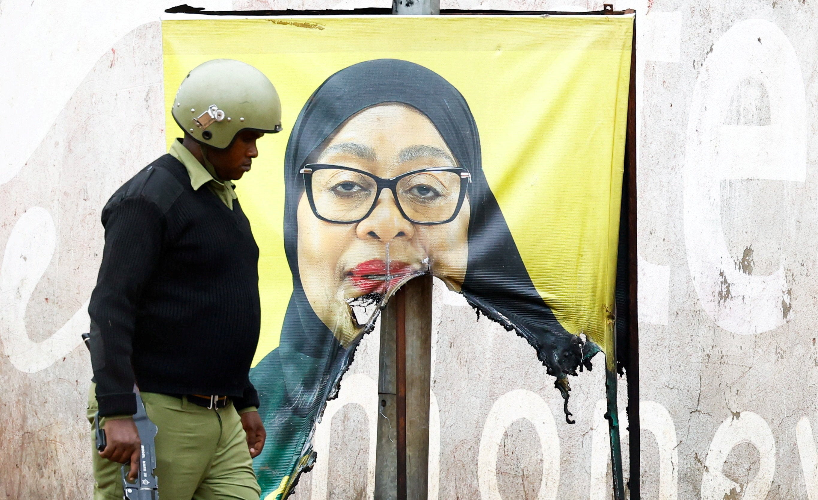 <p>A Tanzanian riot police officer walks past a vandalised campaign poster of President Samia Suluhu Hassan.</p>