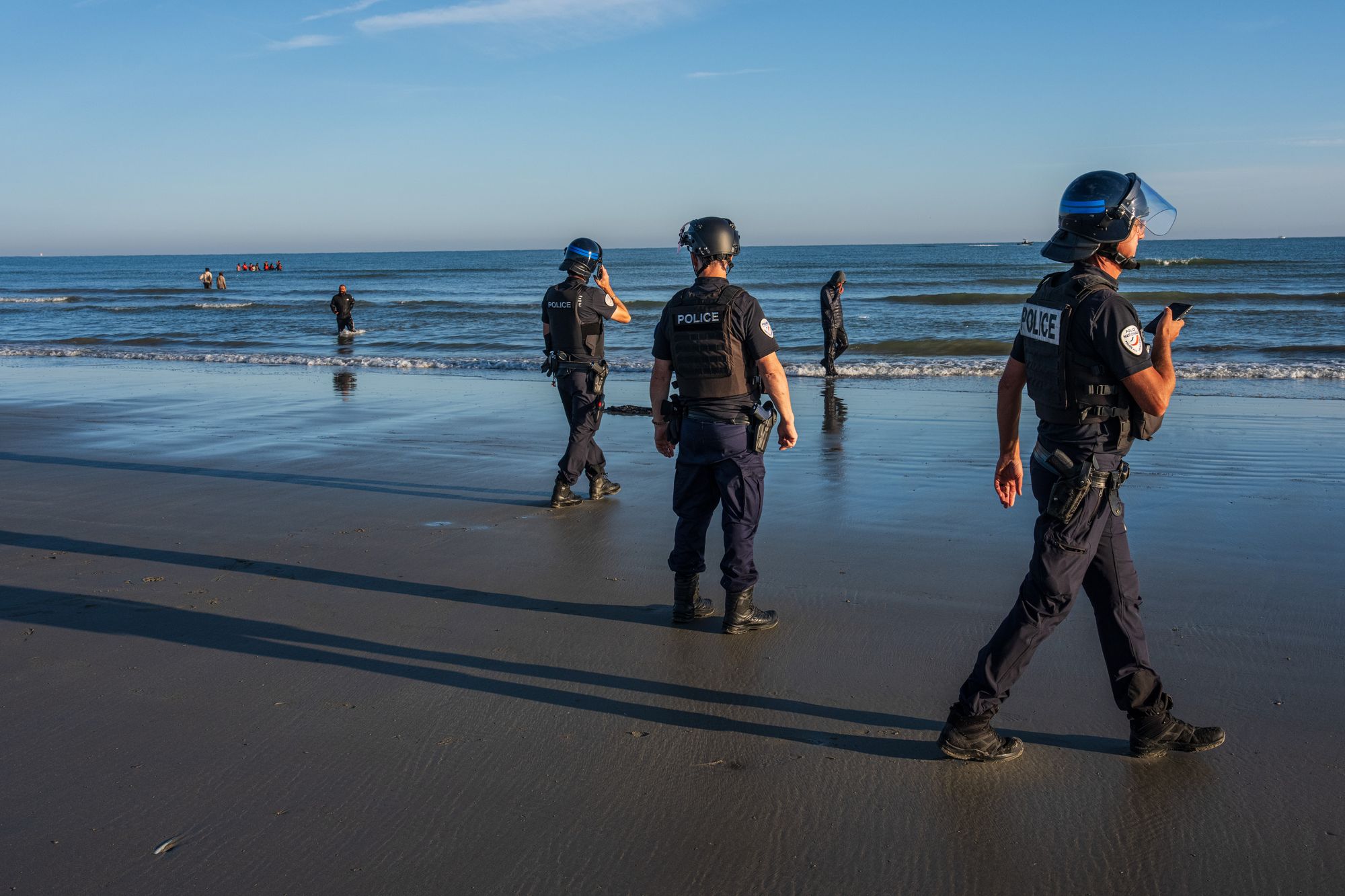 Migrants walk past French police officers after failing to board a dinghy to cross the English Channel on August 25, 2025 in Gravelines, France. French authorities have reportedly agreed to start intercepting dinghies in the Channel.