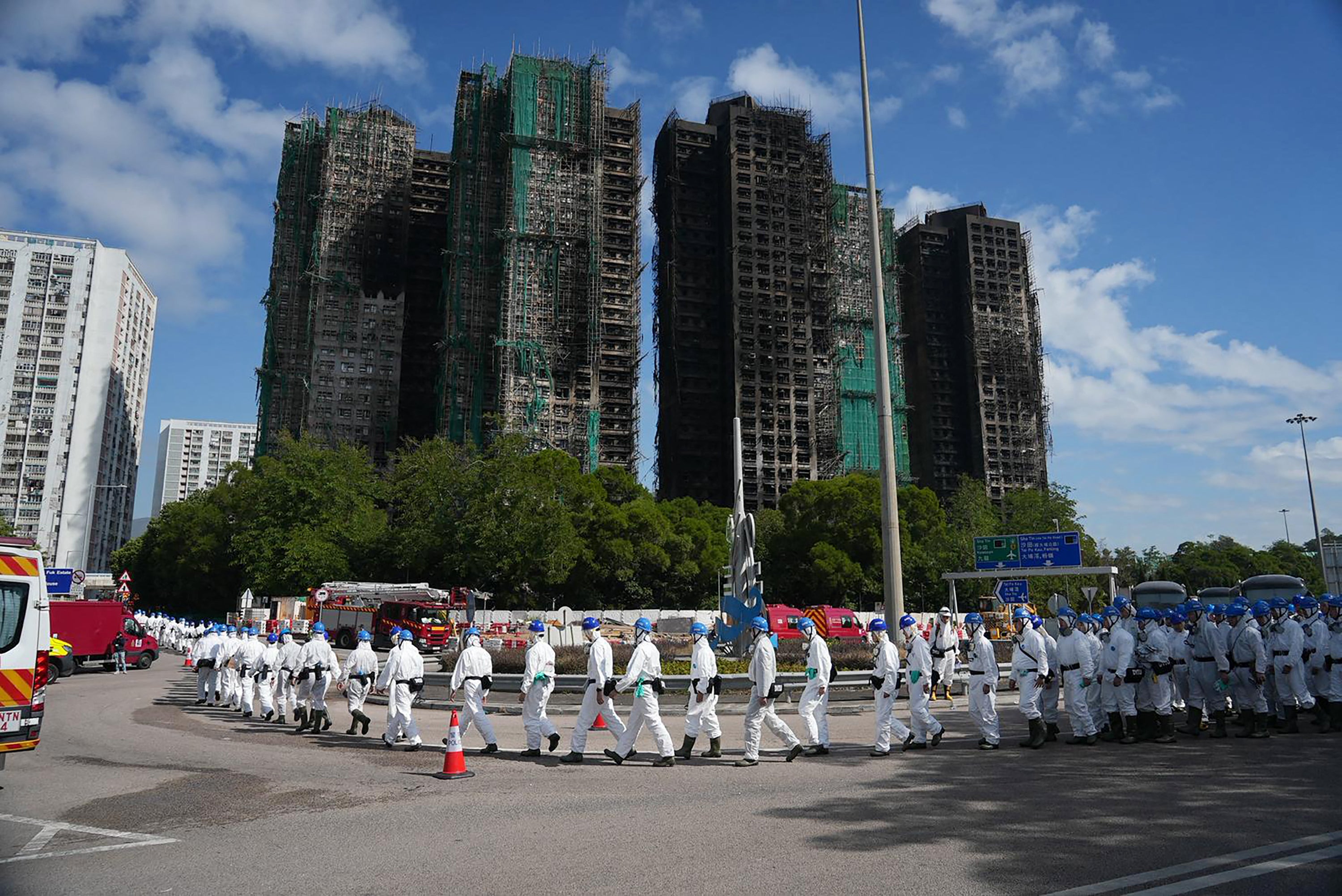 <p>Police officers from the Disaster Victim Identification Unit (DVIU), dressed in white-coloured full-body protective gear, walk past the housing blocks of Wang Fuk Court in the aftermath of the deadly November 26 fire, in Hong Kong</p>