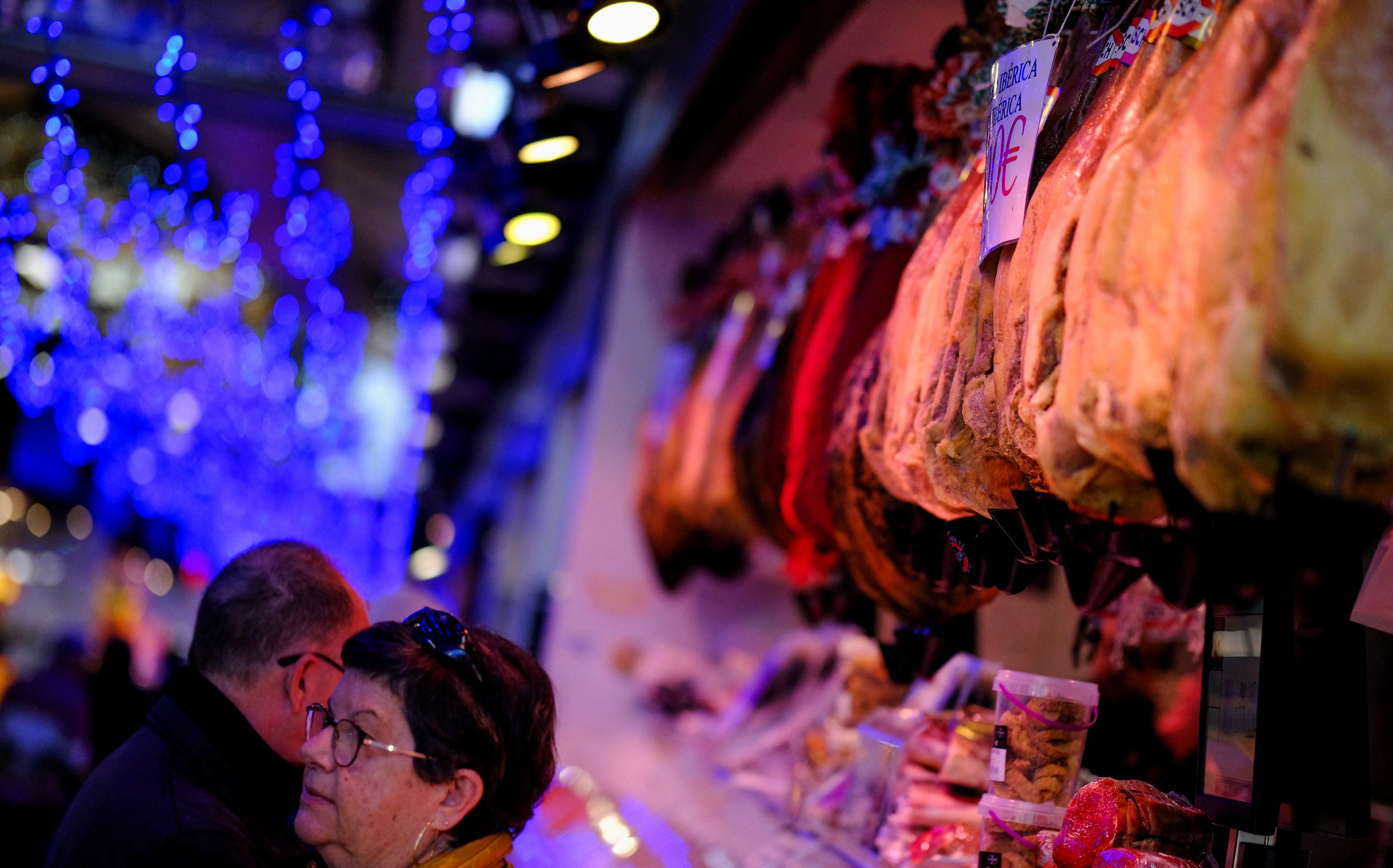 People buy food in front of Spanish Bellota ham hanging at a "Mercado de Abastos" (food market) in Barcelona, Spain