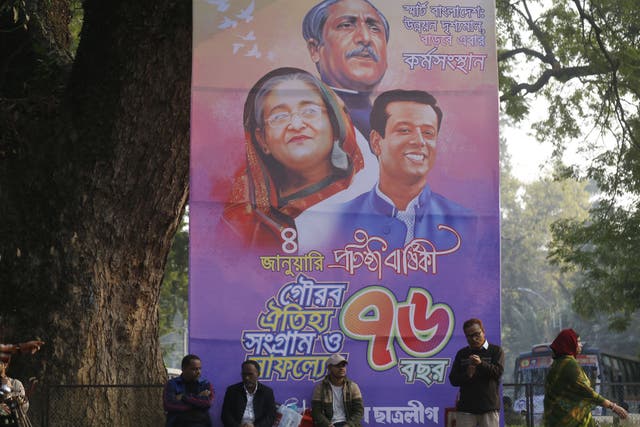<p>People sit next to a banner marking the 74th founding anniversary of Awami League party with images of Sheikh Hasina, her father Sheikh Mujibur Rahman and son Sajeeb Wazed</p>