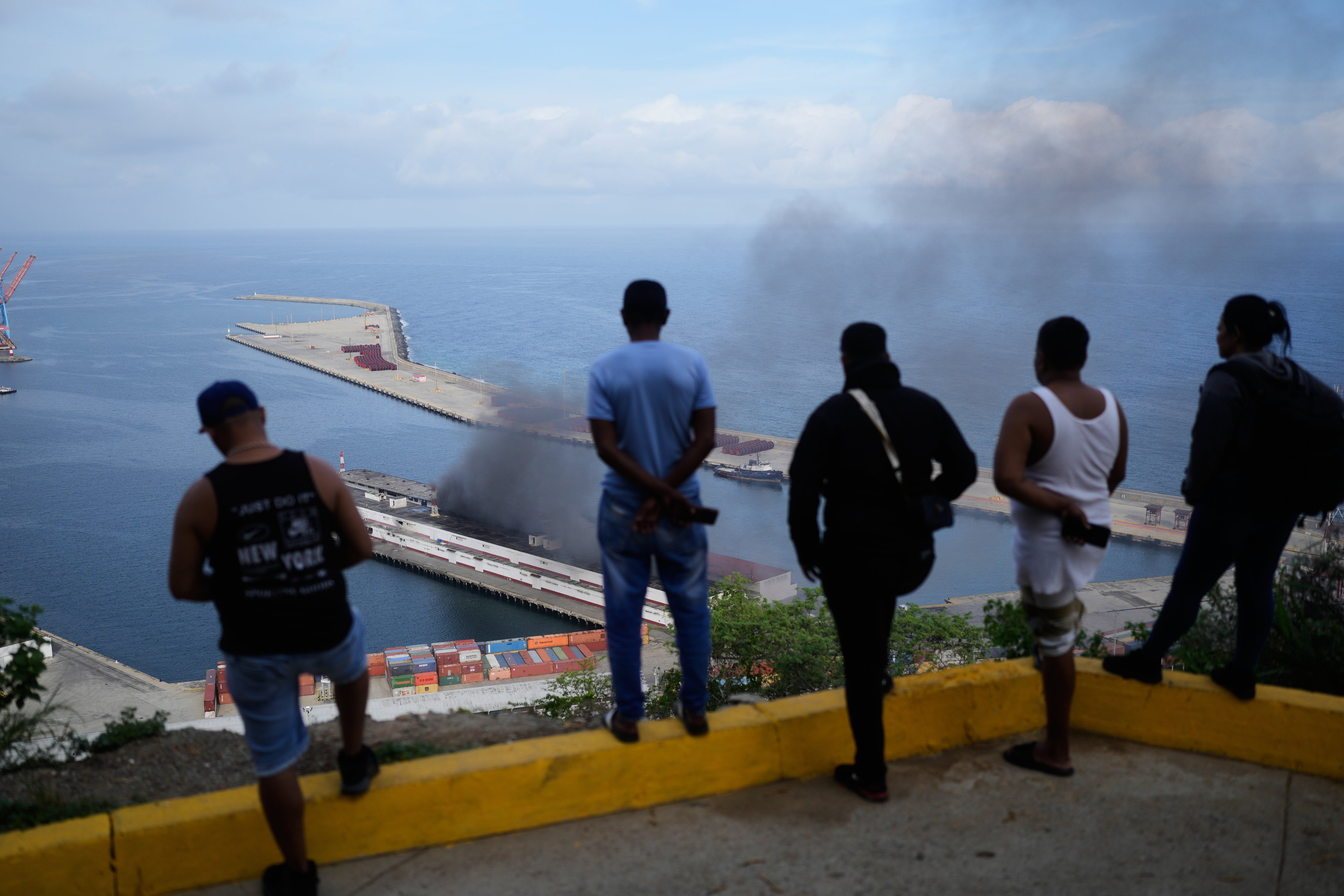Men watch smoke rising from a dock after explosions were heard at La Guaira port, Venezuela, Saturday, Jan. 3, 2026. (AP Photo/Matias Delacroix) Venezuela US