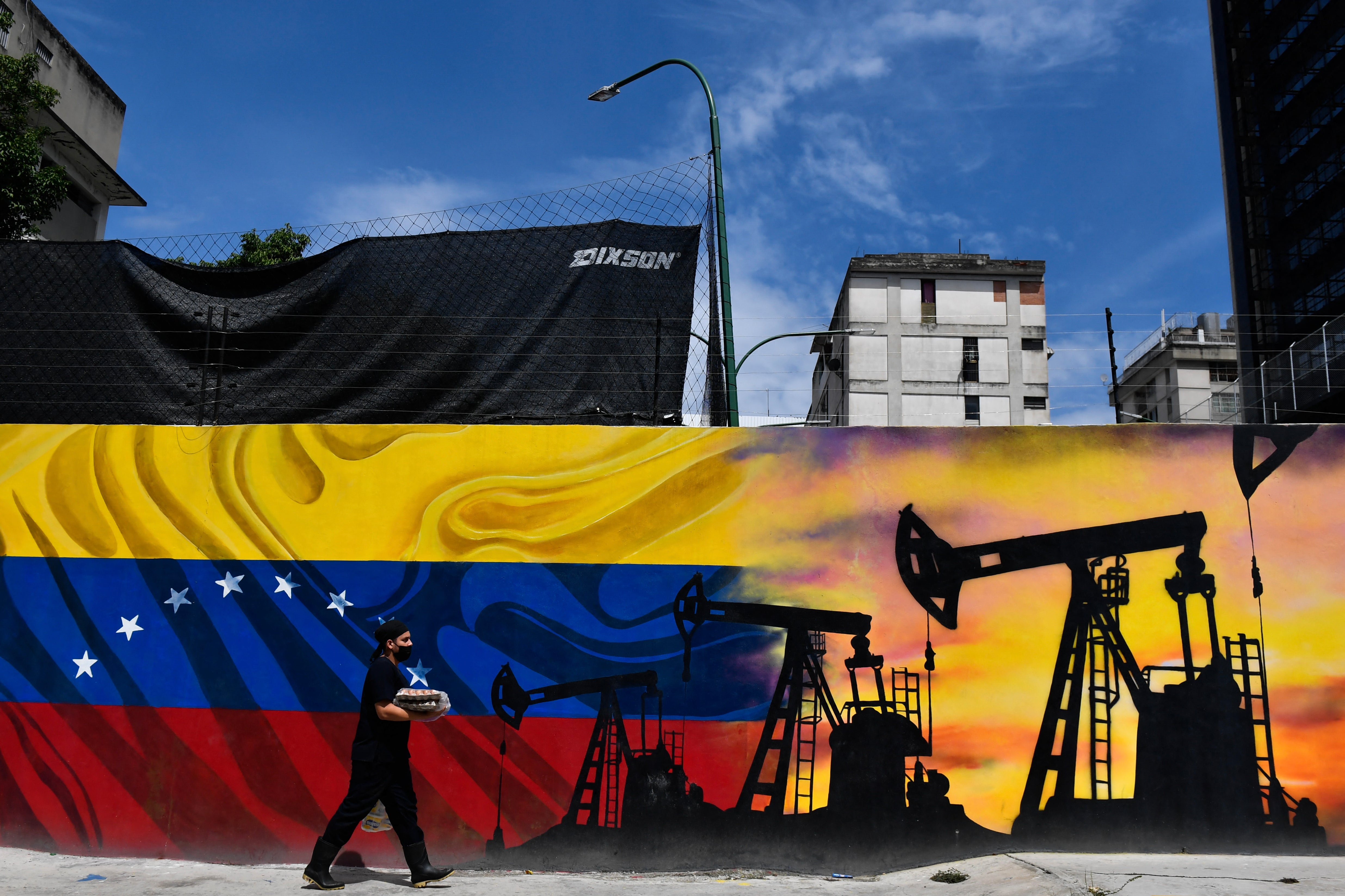 A man wearing a face masks walks past a mural depicting an oil pump and the Venezuelan flag in a street of Caracas, on May 26, 2022
