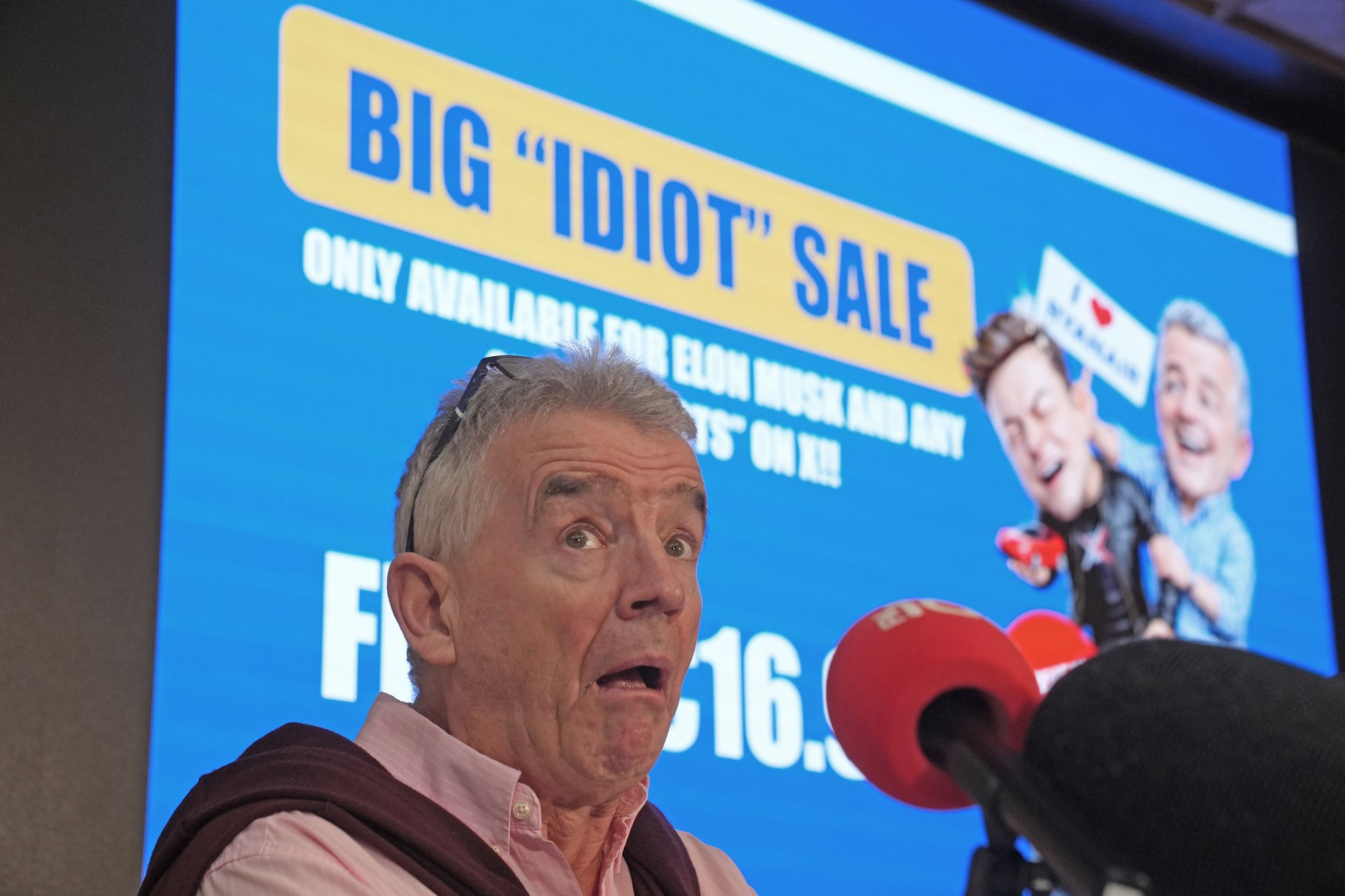 <p>Ryanair Group CEO Michael O'Leary in front of his ‘big “idiot” sale’ press conference screen showing a cartoon Musk and O’Leary</p>