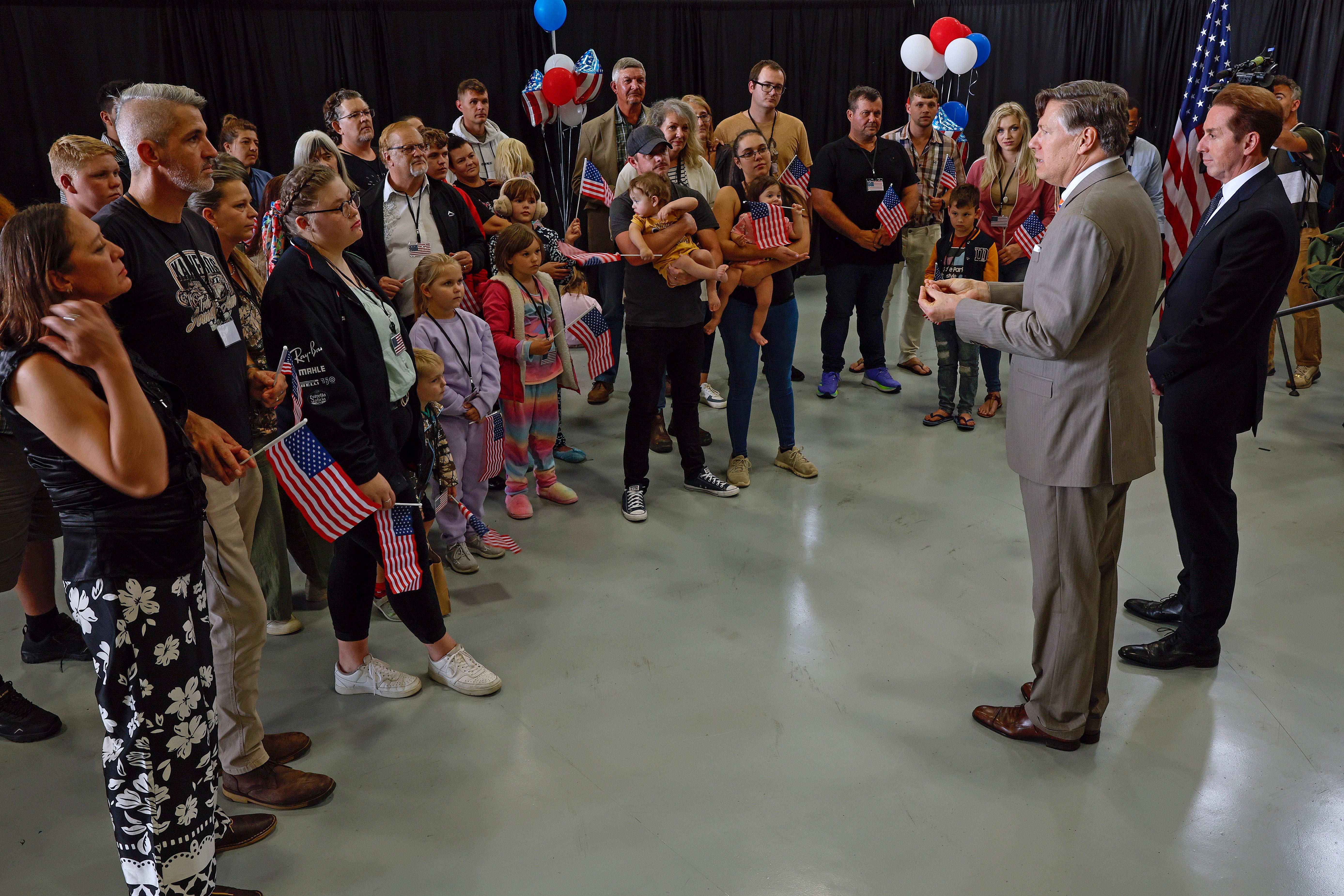Newly arrived South Africans listen to U.S. Deputy Secretary of State Christopher Landau and Homeland Security Deputy Secretary Troy Edgar (R) deliver welcome statements in a hangar at Atlantic Aviation Dulles near Washington Dulles International Airport on May 12