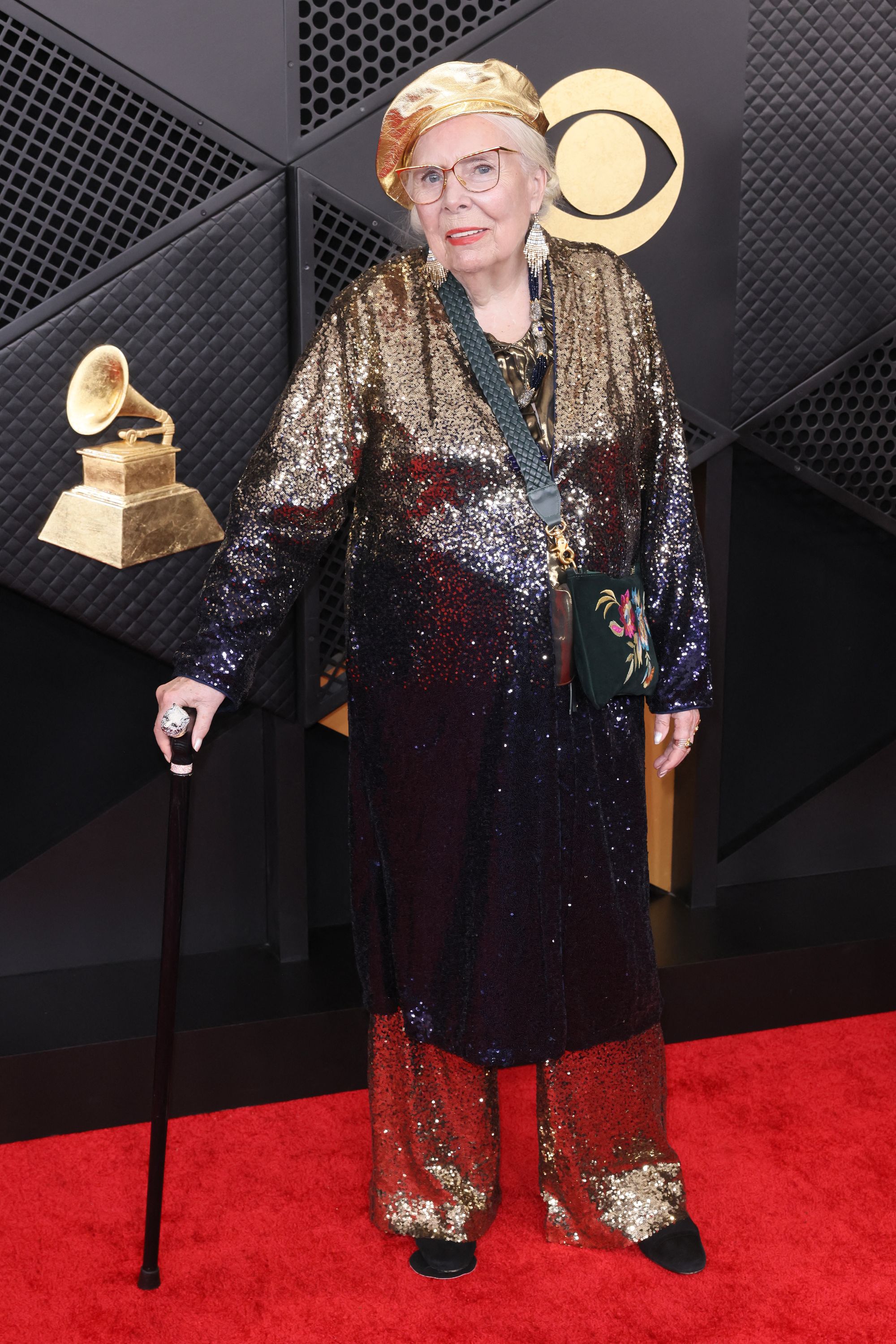 Joni Mitchell took over the red carpet in all sparkles, wearing gold flare pants and a matching long-sleeved dress. She paired the look with a black floral cross-body purse