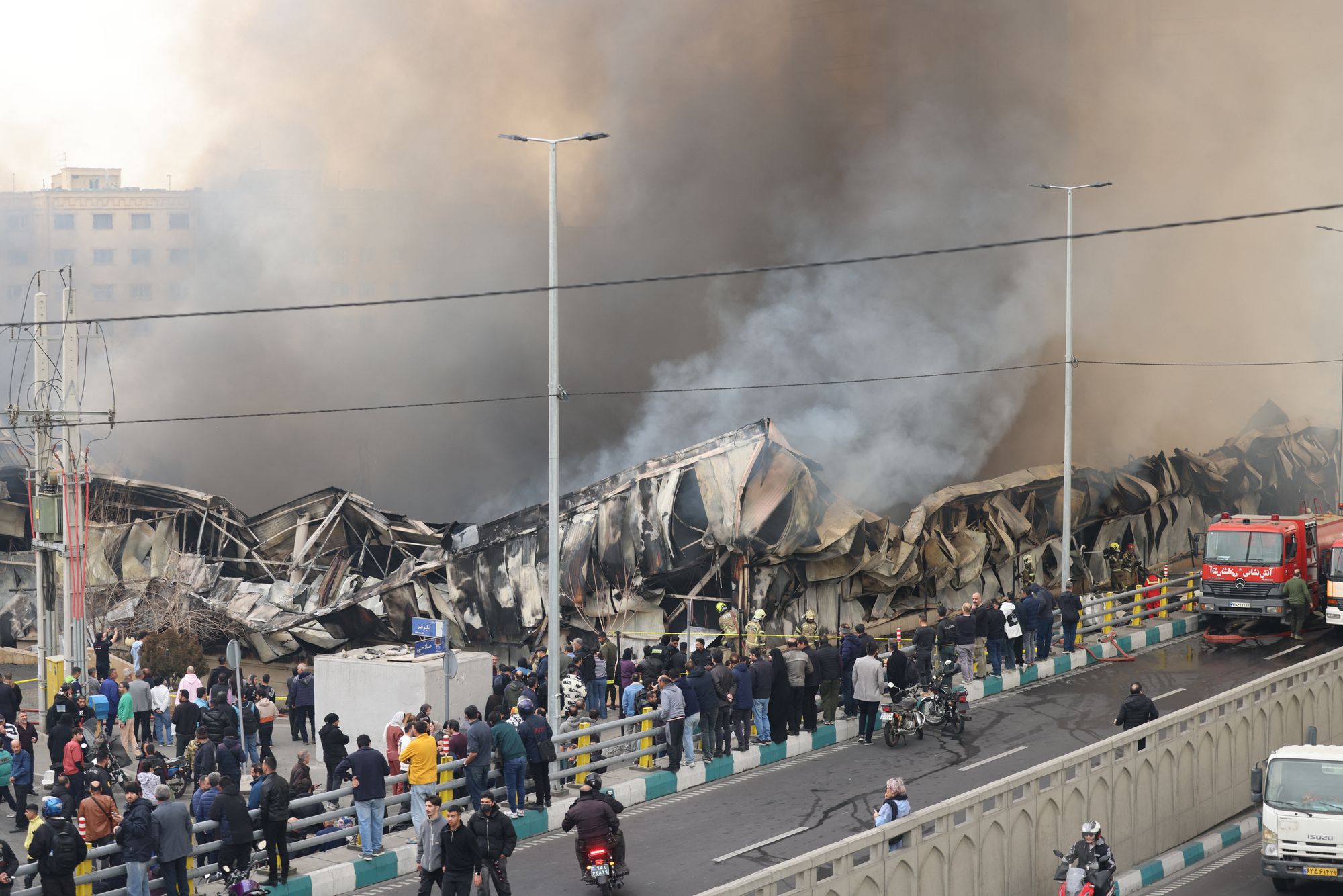 People stand and watch as smoke billows from a fire that broke out in Jannat Bazaar, west of Tehran, on 3 February 2026.