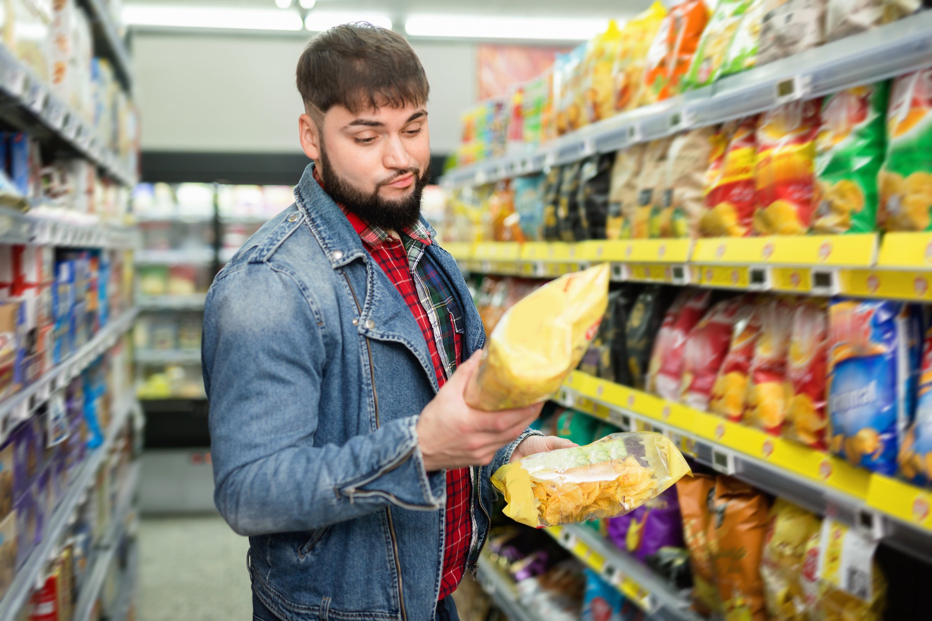 Portrait of doubting smiling glad friendly bearded guy choosing potato chips at grocery store