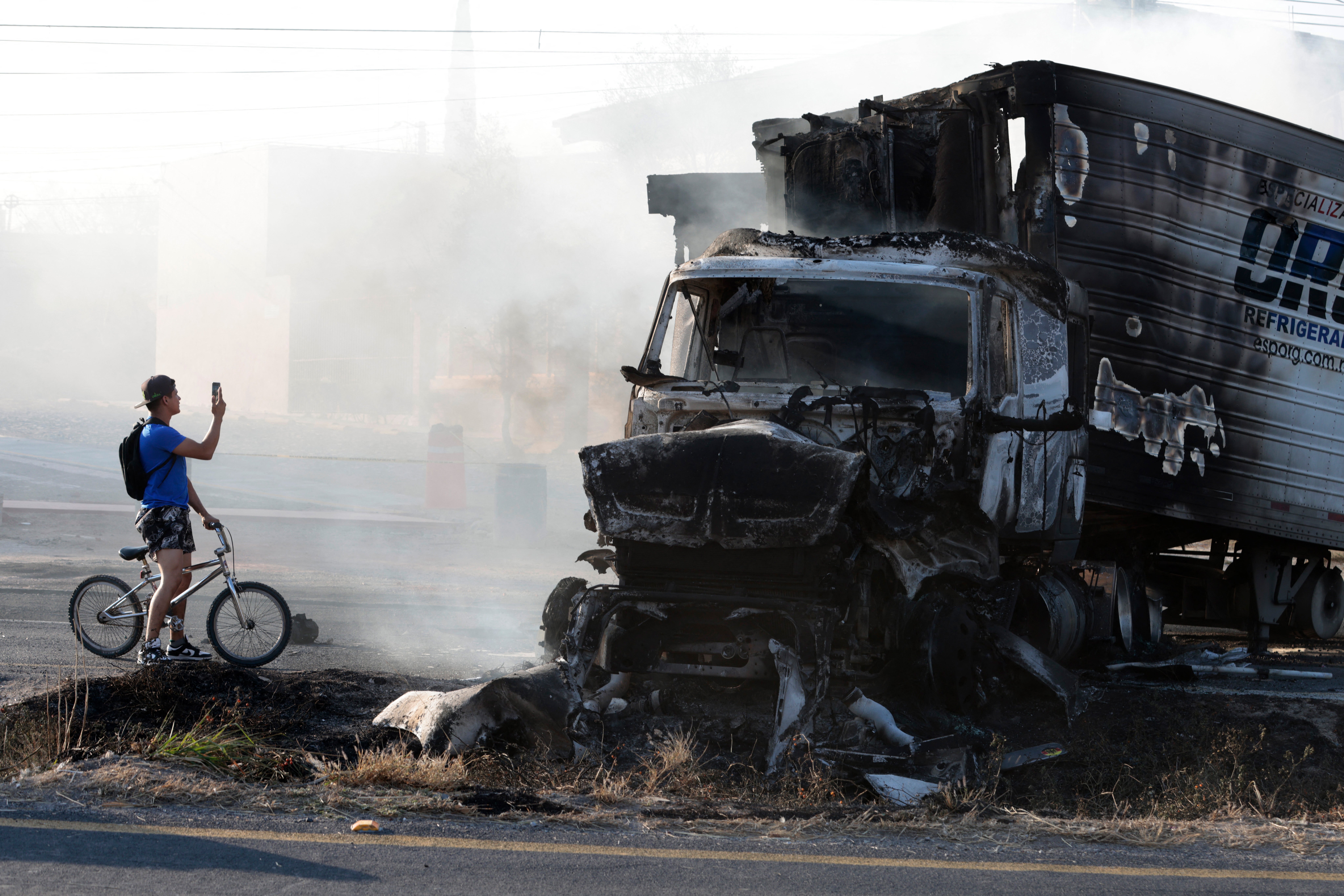 <p>A man takes a photo of a burned truck, allegedly set on fire by organised crime groups in response to an operation to arrest a high-priority security target, on a highway near Acatlan de Juarez, Jalisco, Mexico, on February 22, 2026</p>