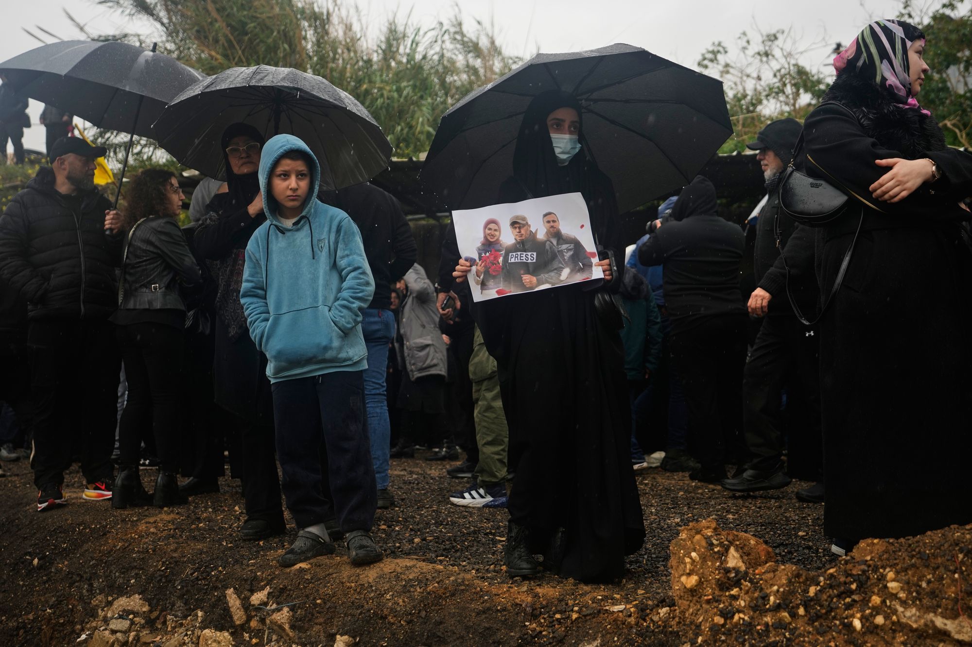 A woman holds a poster showing Hezbollah's Al-Manar TV correspondent Ali Shoeib, center, Al-Mayadeen TV reporter Fatima Ftouni, left, and cameraman Ali Ftouni as mourners gather near a freshly dug grave during their funeral at a temporary cemetery in Dahiyeh, Beirut's southern suburbs, Beirut, Lebanon, Sunday, March 29, 2026. (AP Photo/Hassan Ammar)