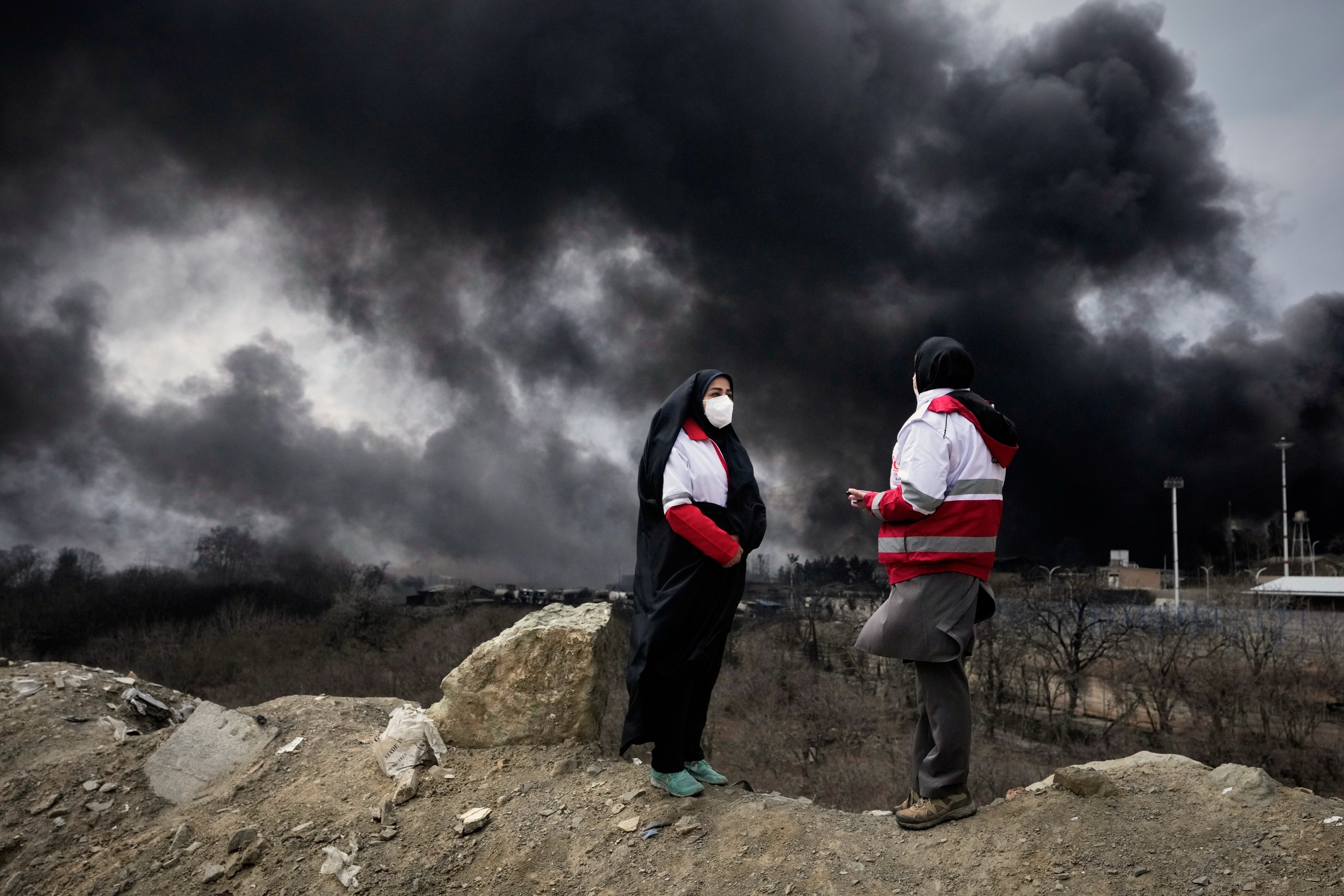 Two women from the Iranian Red Crescent Society stand as a thick plume of smoke from a U.S.-Israeli strike on an oil storage facility late Saturday rises into the sky in Tehran, Iran, Sunday, March 8, 2026. (AP Photo/Vahid Salemi, File)
