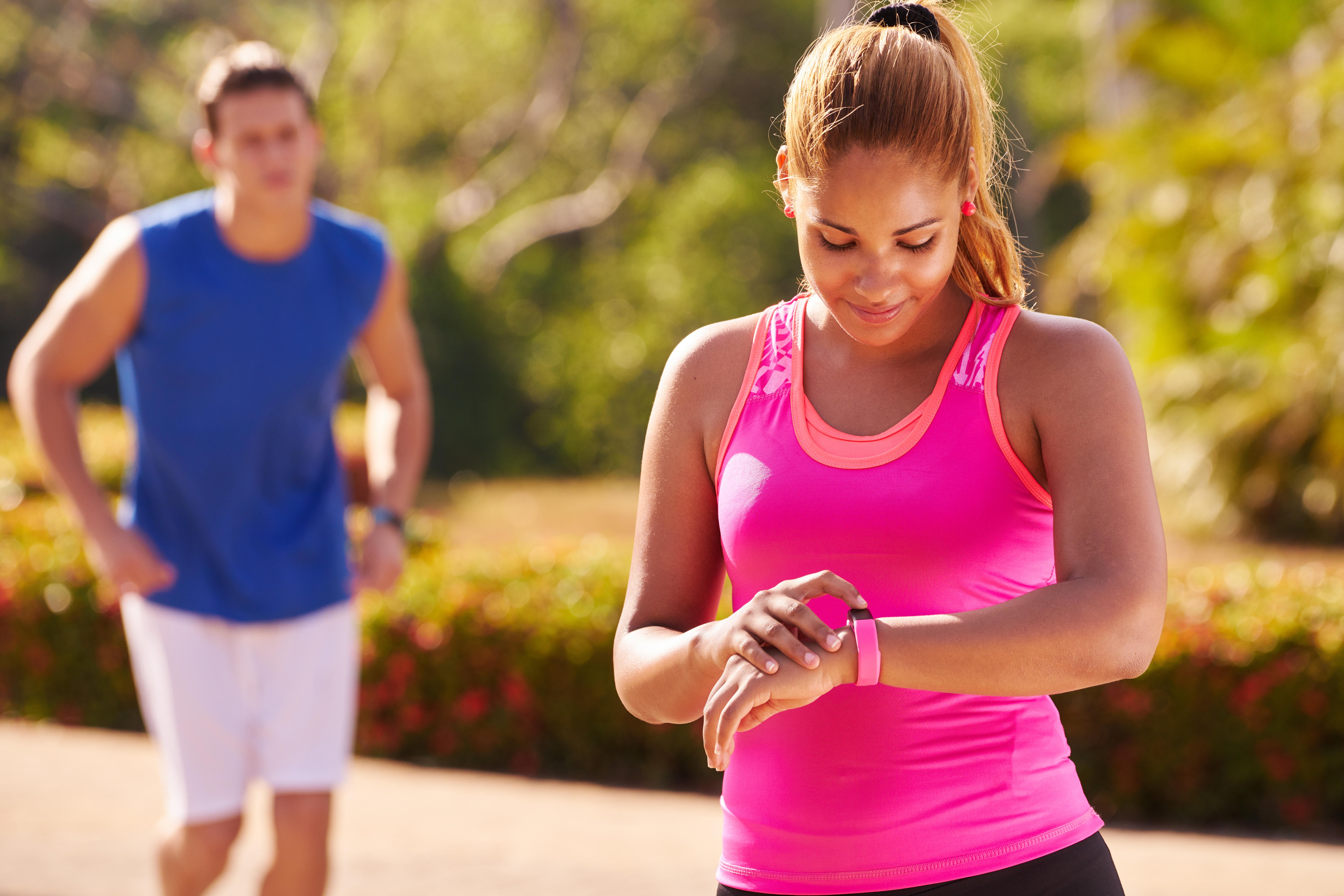 A woman wearing a pink sports top looking at her smartwatch after a run, and a man running behind her in the background (Alamy/PA)