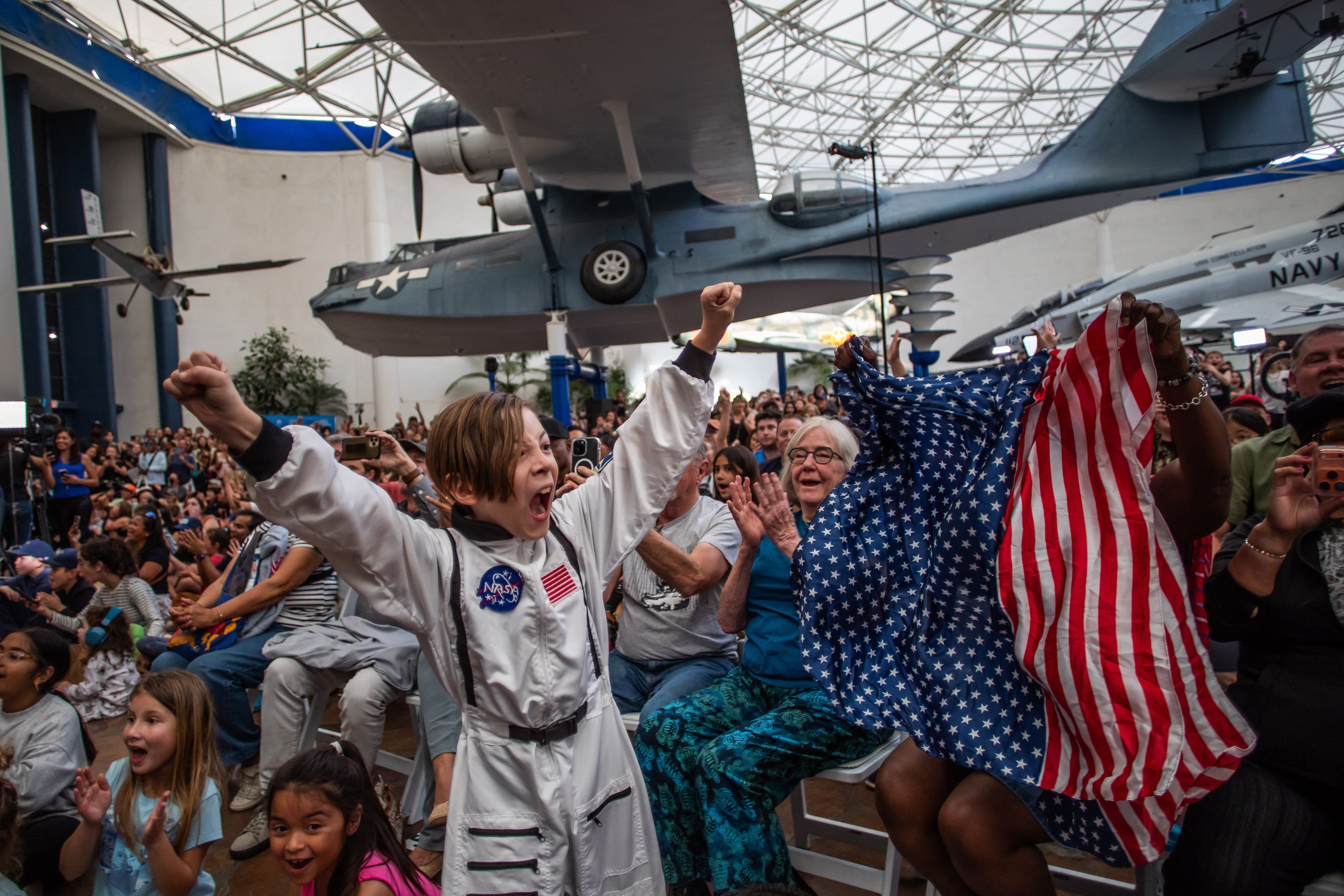 A young boy wearing an astronaut costume cheers next to a woman waving a flag as they watch a live broadcast of the return of the Artemis II crew members to Earth at the San Diego Air and Space Museum during a watch party for the crew's splash down in the Pacific Ocean, in San Diego, California, on April 10, 2026.