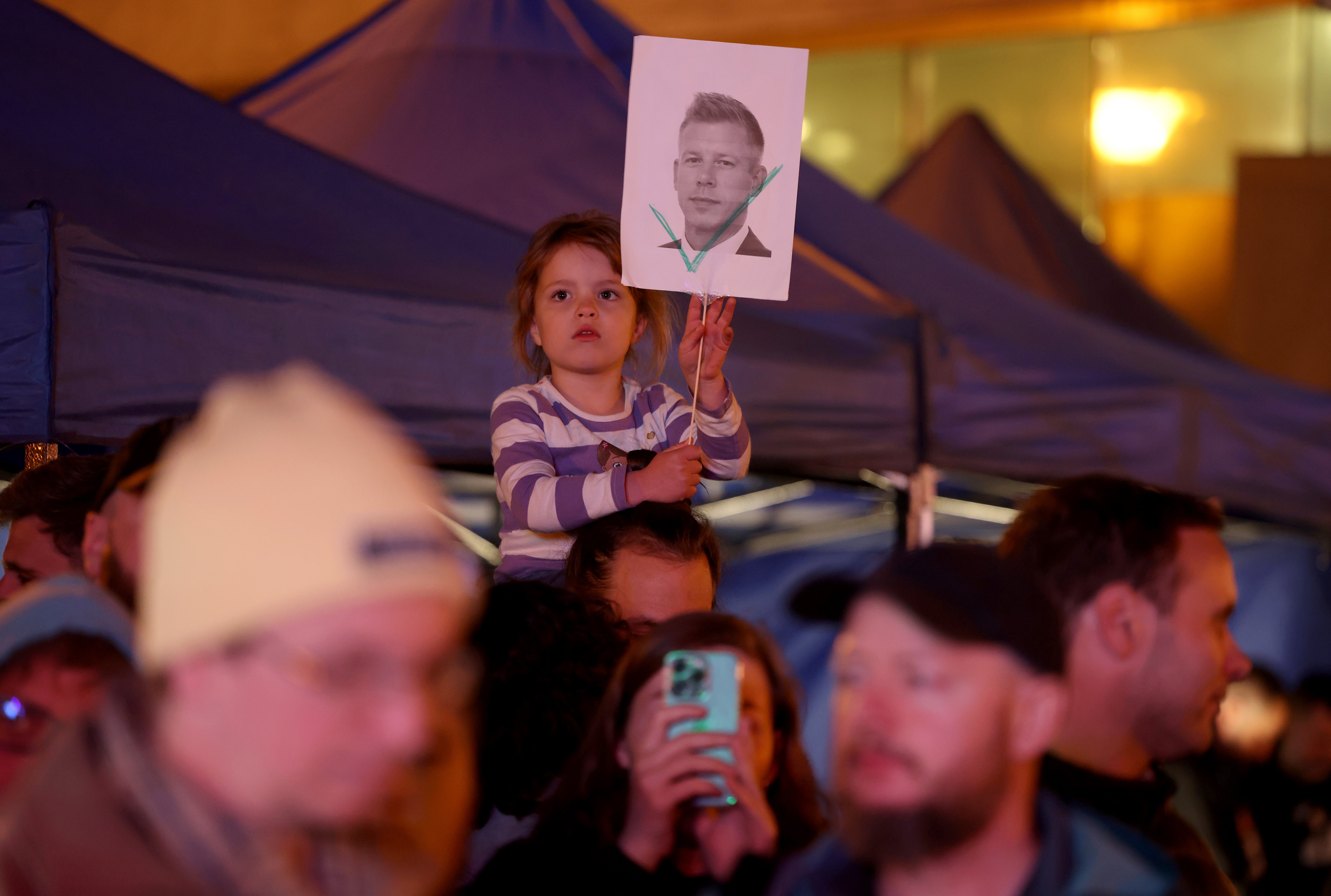 A child holds a picture of opposition candidate Peter Magyar with a green check mark among people gathered at a local outdoor post-election watch party following the closing of polling stations in Hungarian parliamentary elections