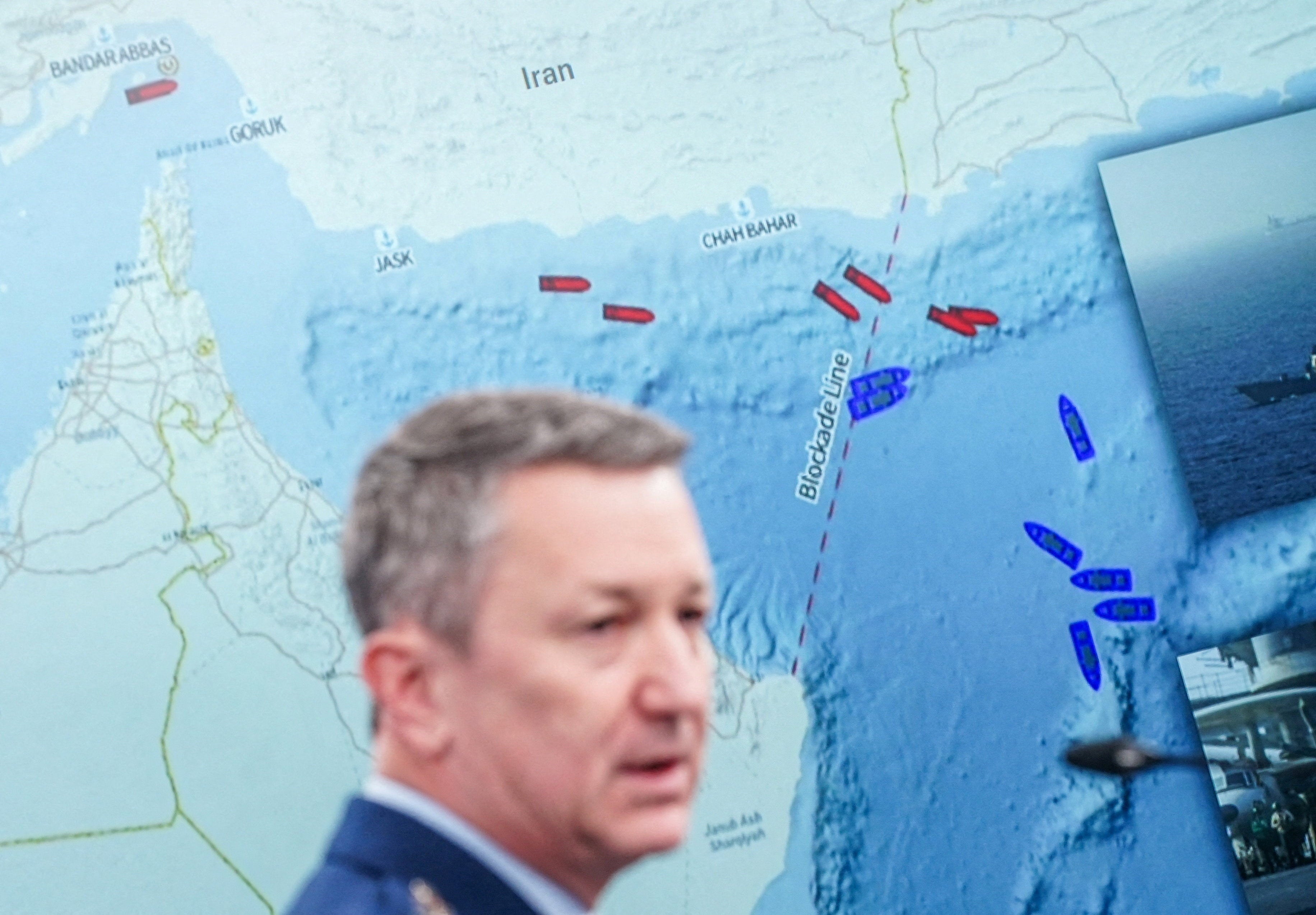 General Dan Caine speaks during a briefing on the Iran war, with a map in the background showing a blockade line on the Strait of Hormuz