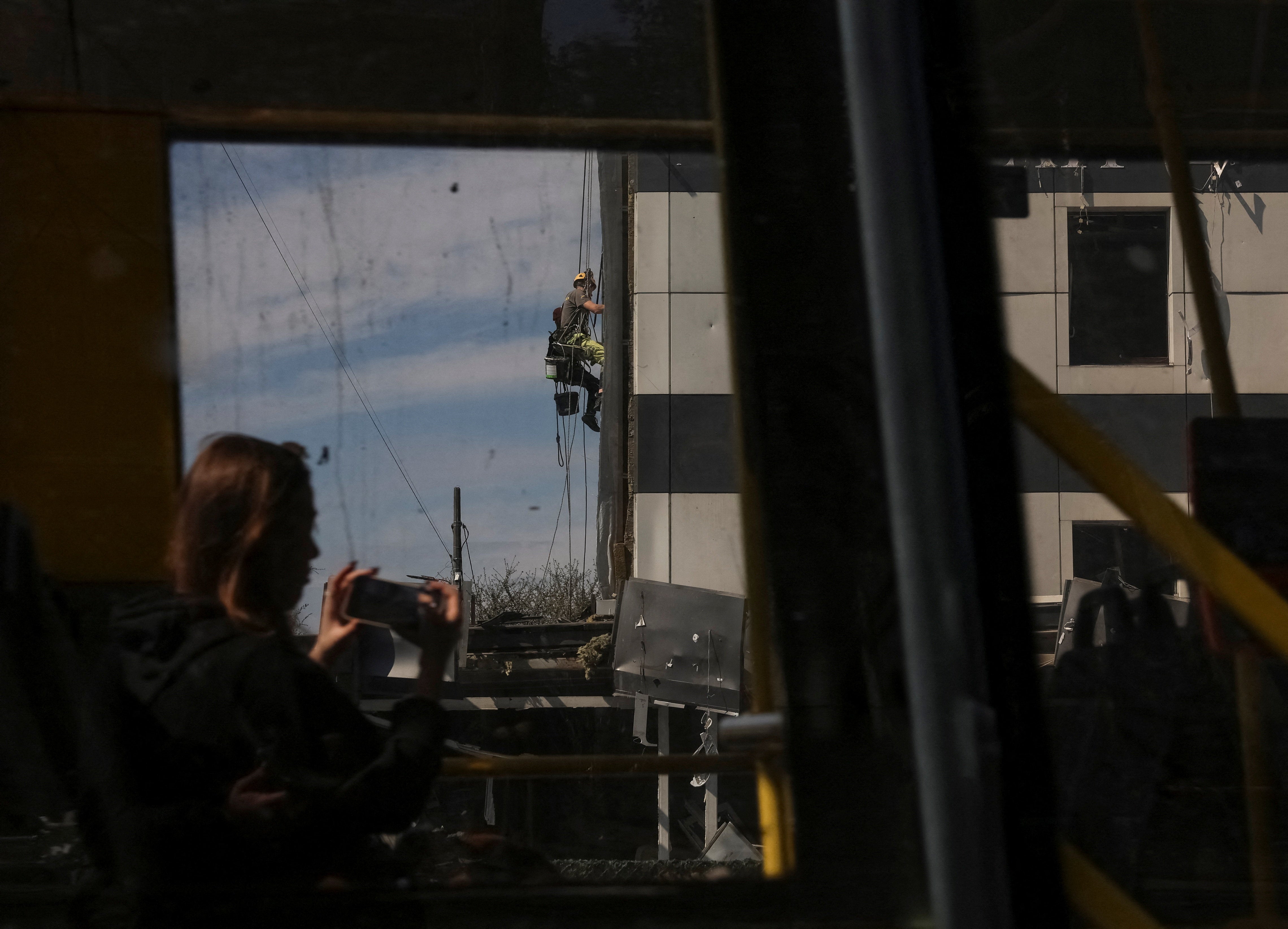 A woman rides a bus and takes pictures as a worker installs an OSB board in place of a broken window of a hotel hit by a yesterday's Russian missile and drone strike, amid Russia's attack on Ukraine, in Kyiv, Ukraine April 17,