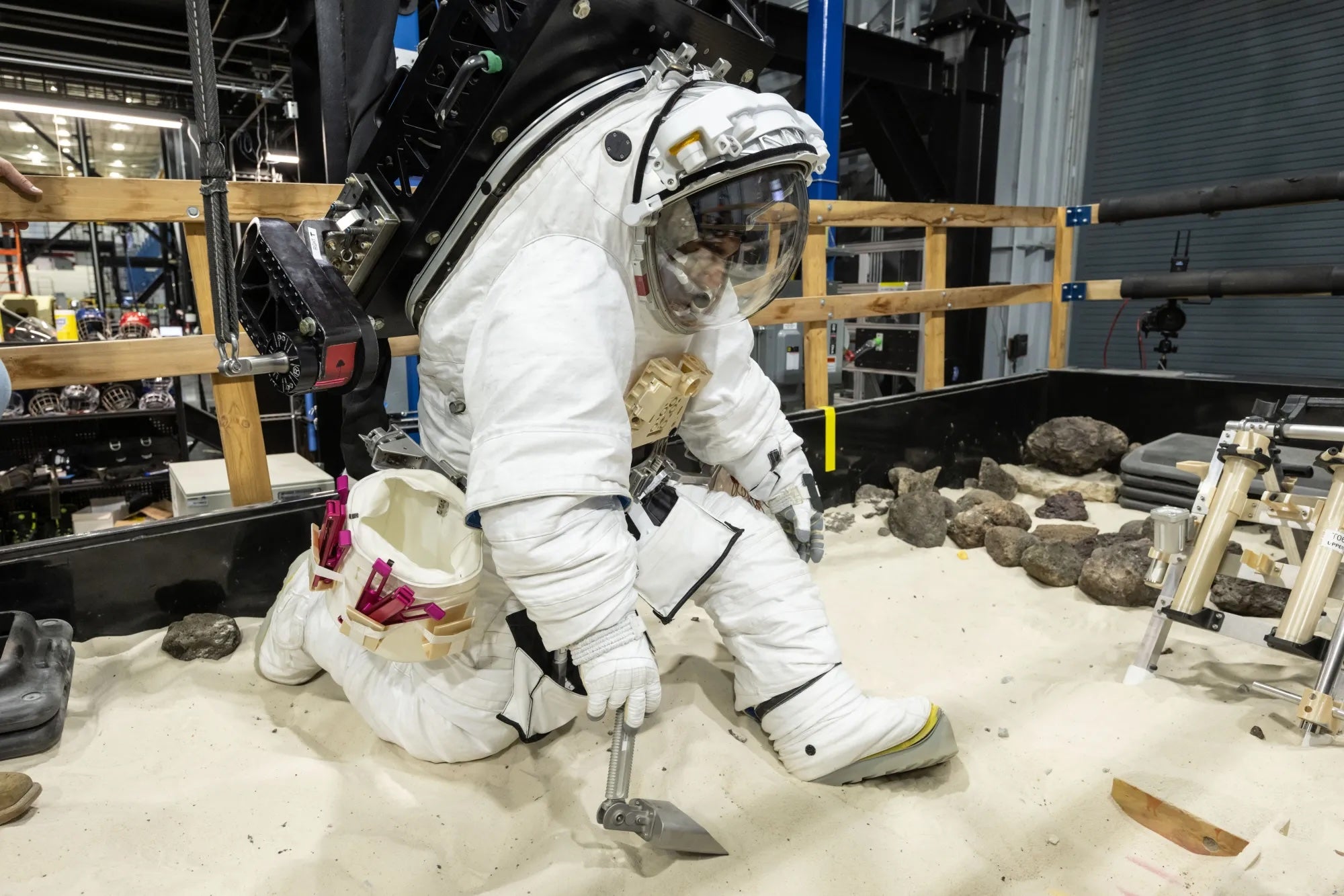 A NASA crew member practices using lunar tools to collect geology samples at NASA’s Johnson Space Center during a pressure test in an Artemis III lunar spacesuit developed by Axiom Space
