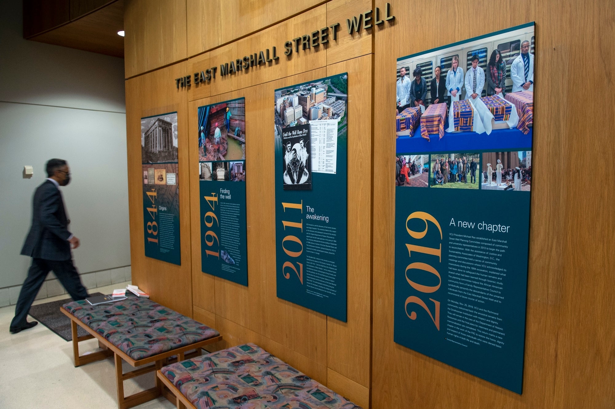 In this photo provided by Virginia Commonwealth University, a man walks past a wall display about the effort to identify and honor the dozens of people whose remains were found in an abandoned well on the VCU campus in Richmond, Va., in 2021. (VCU via AP)