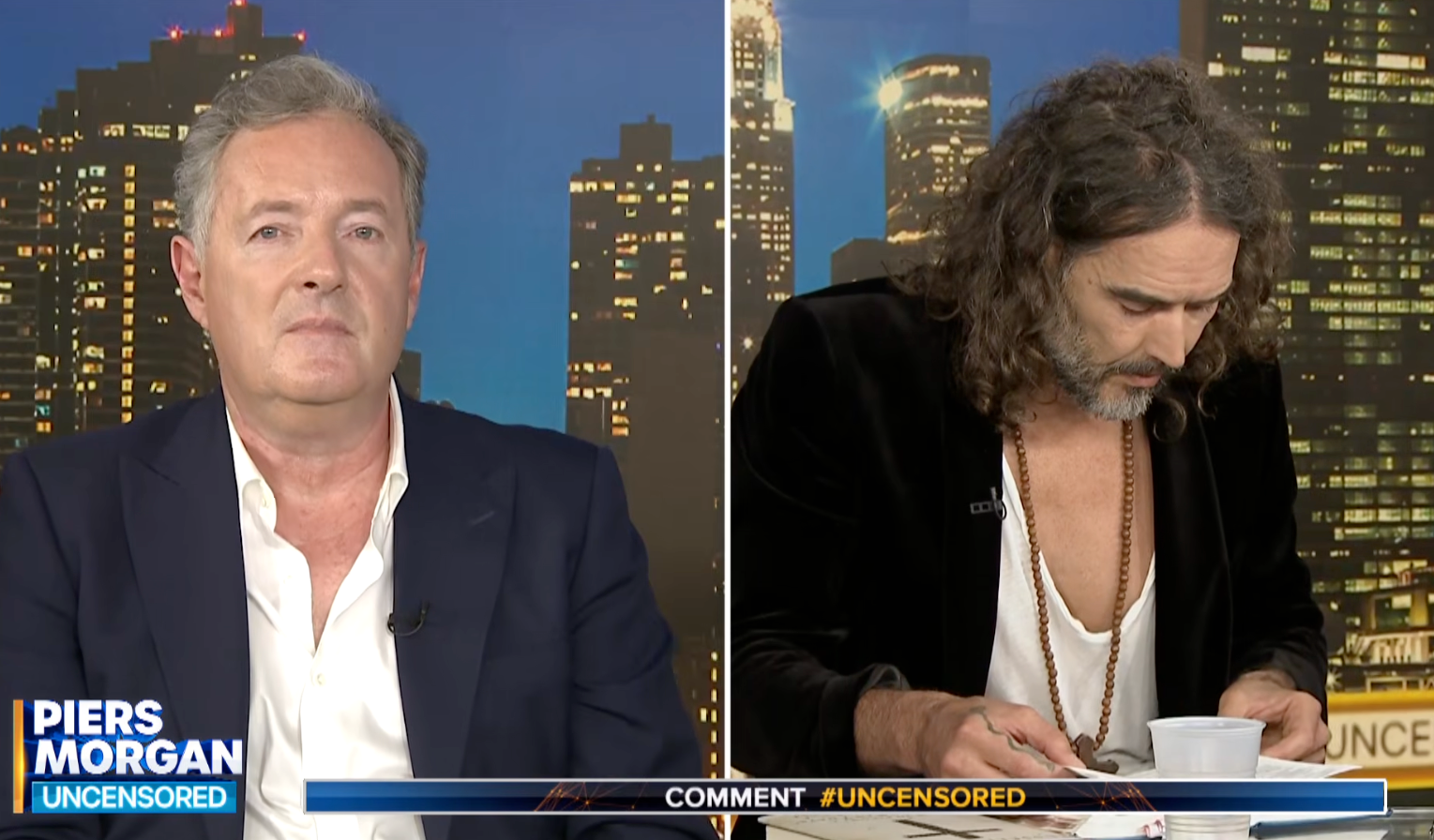 Piers Morgan awkwardly looks at the camera as Russell Brand struggles to find a Bible passage
