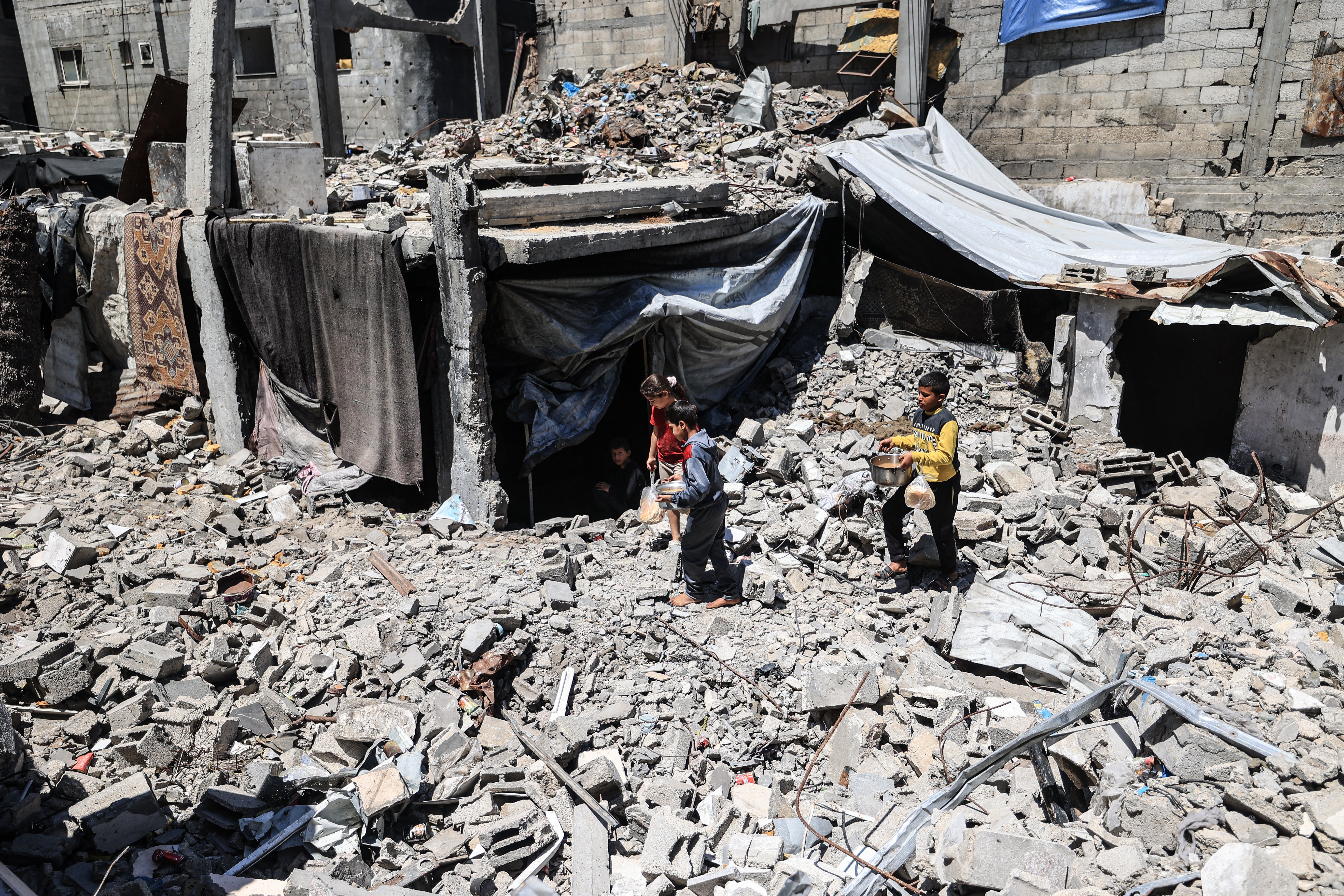 Displaced Palestinian children carry pots of food collected from a charity kitchen to their family shelter beneath the rubble of buildings destroyed by the Israeli military, in the Bureij refugee camp in the central Gaza Strip on May 1