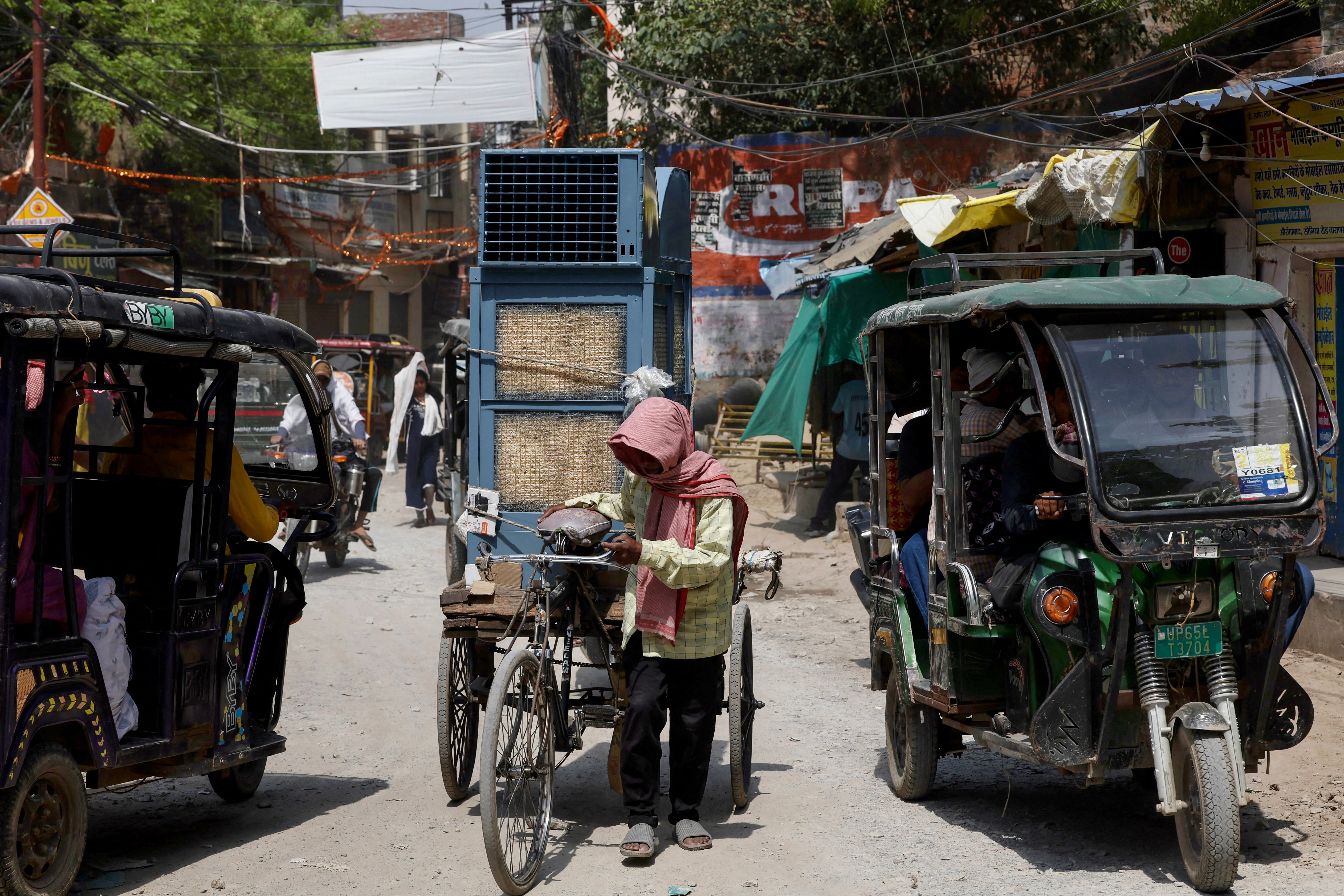 A man with his head covered with a cloth transports a cooler on a cycle cart along a street, on a hot summer day