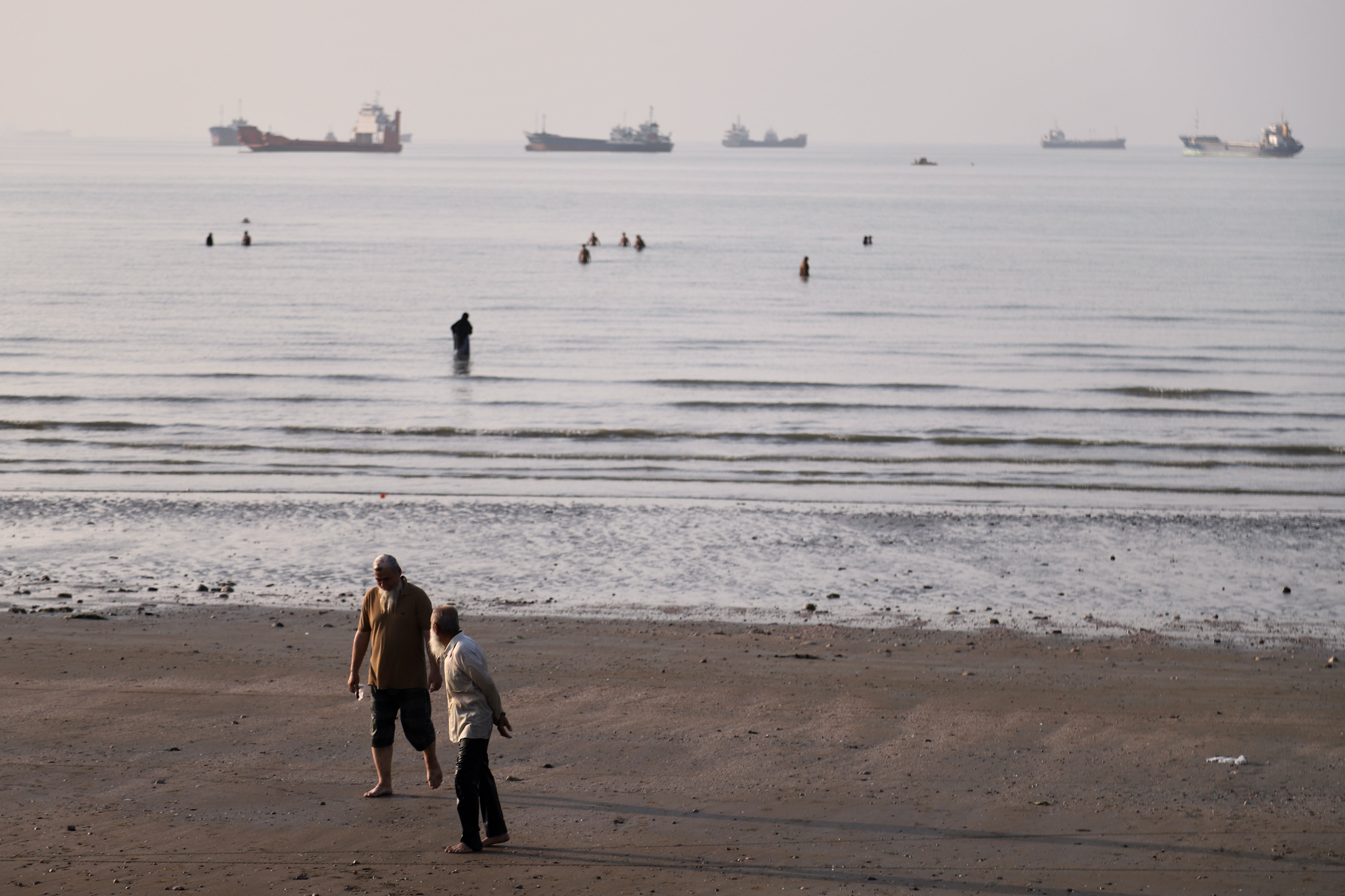 Men walk on the shoreline as people wade in the water in the Strait of Hormuz, where cargo and service vessels are anchored off Bandar Abbas, Iran