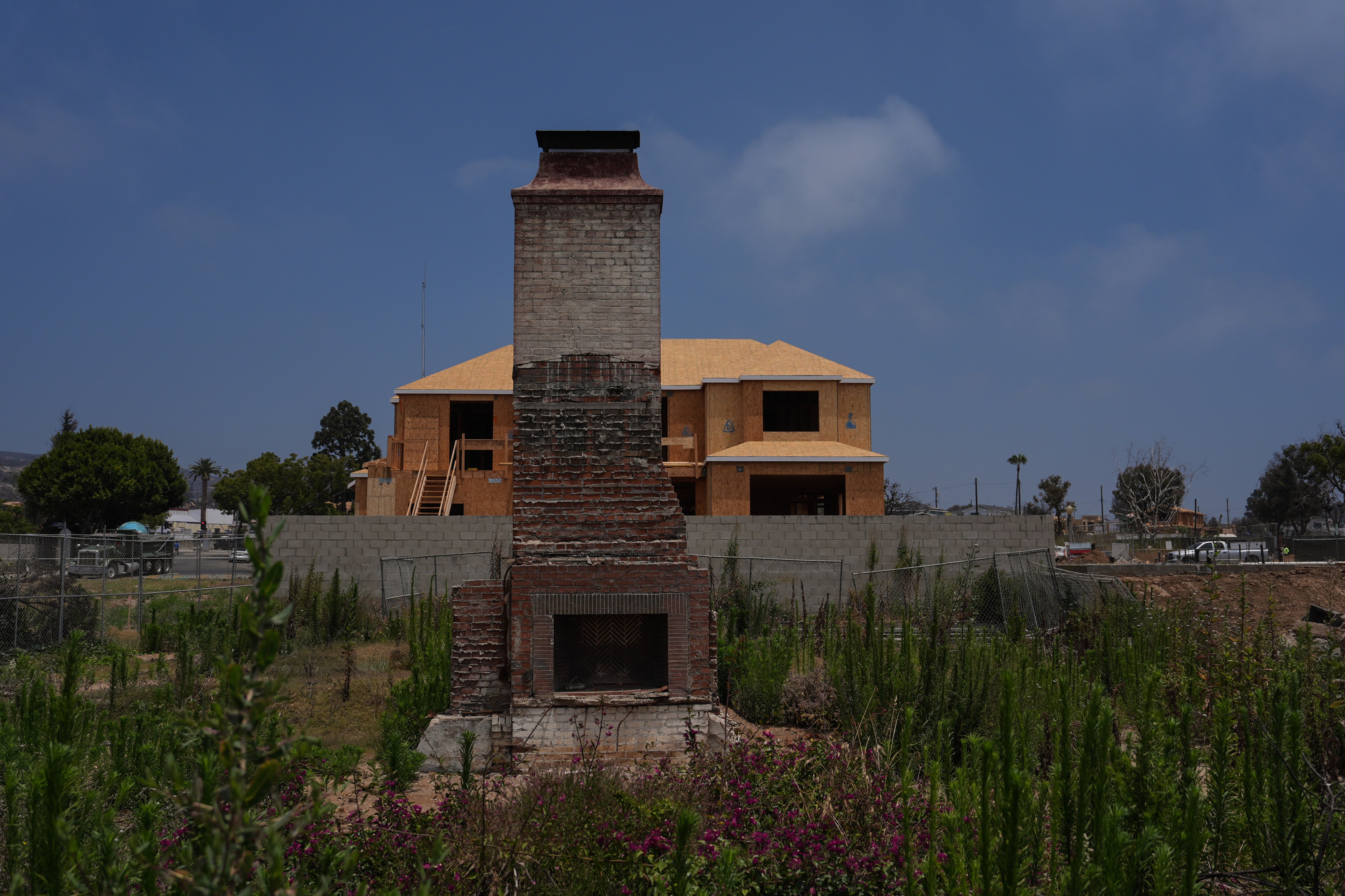 A chimney stands on a lot covered with weeds and wildflowers in front of a home under construction more than a year after the Palisades Fire