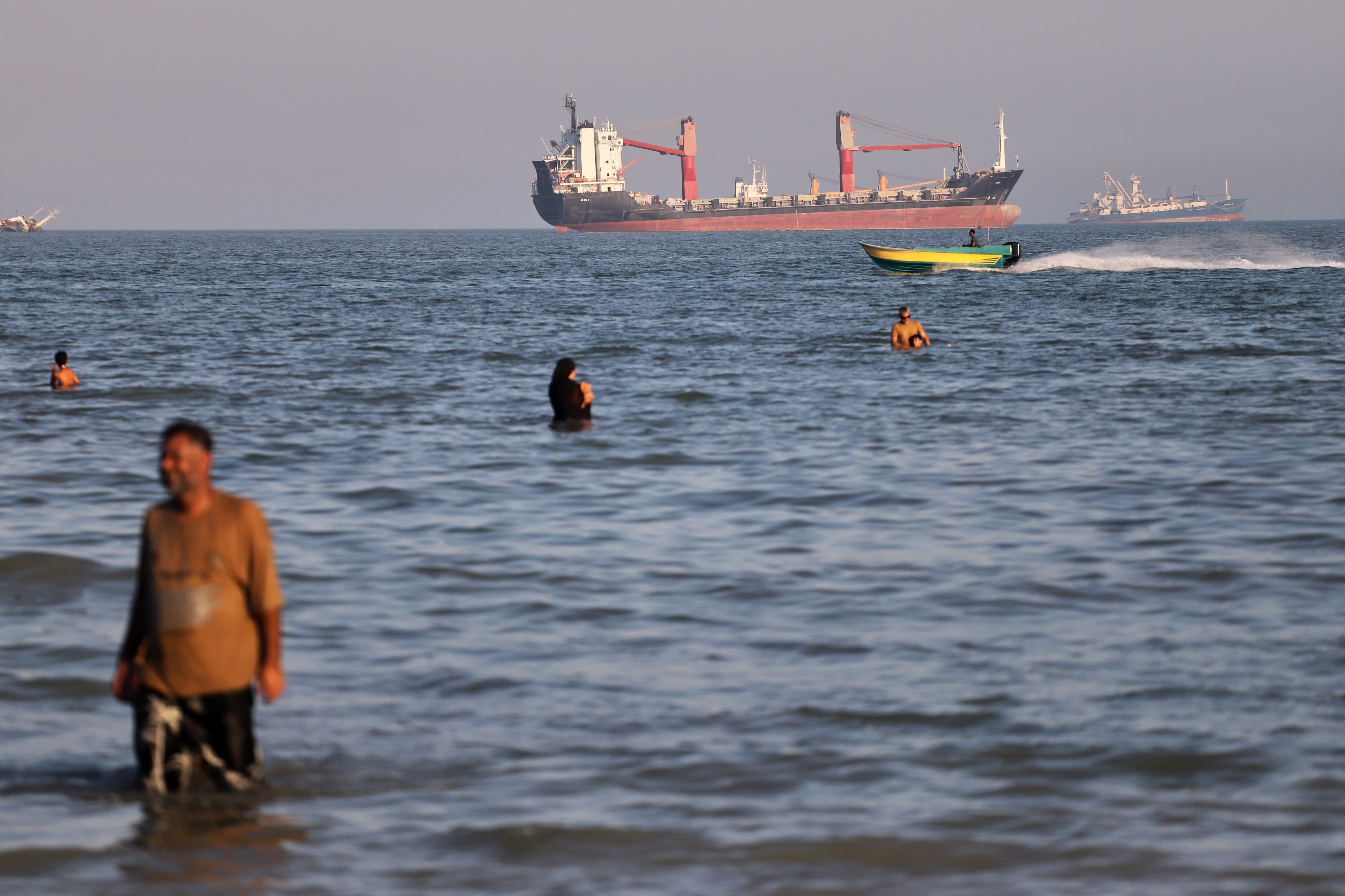 Residents swim in the waters of the Strait of Hormuz as a small motorboat passes cargo ships and other commercial vessels offshore near Bandar Abbas, Iran, Wednesday, 17 June 2026