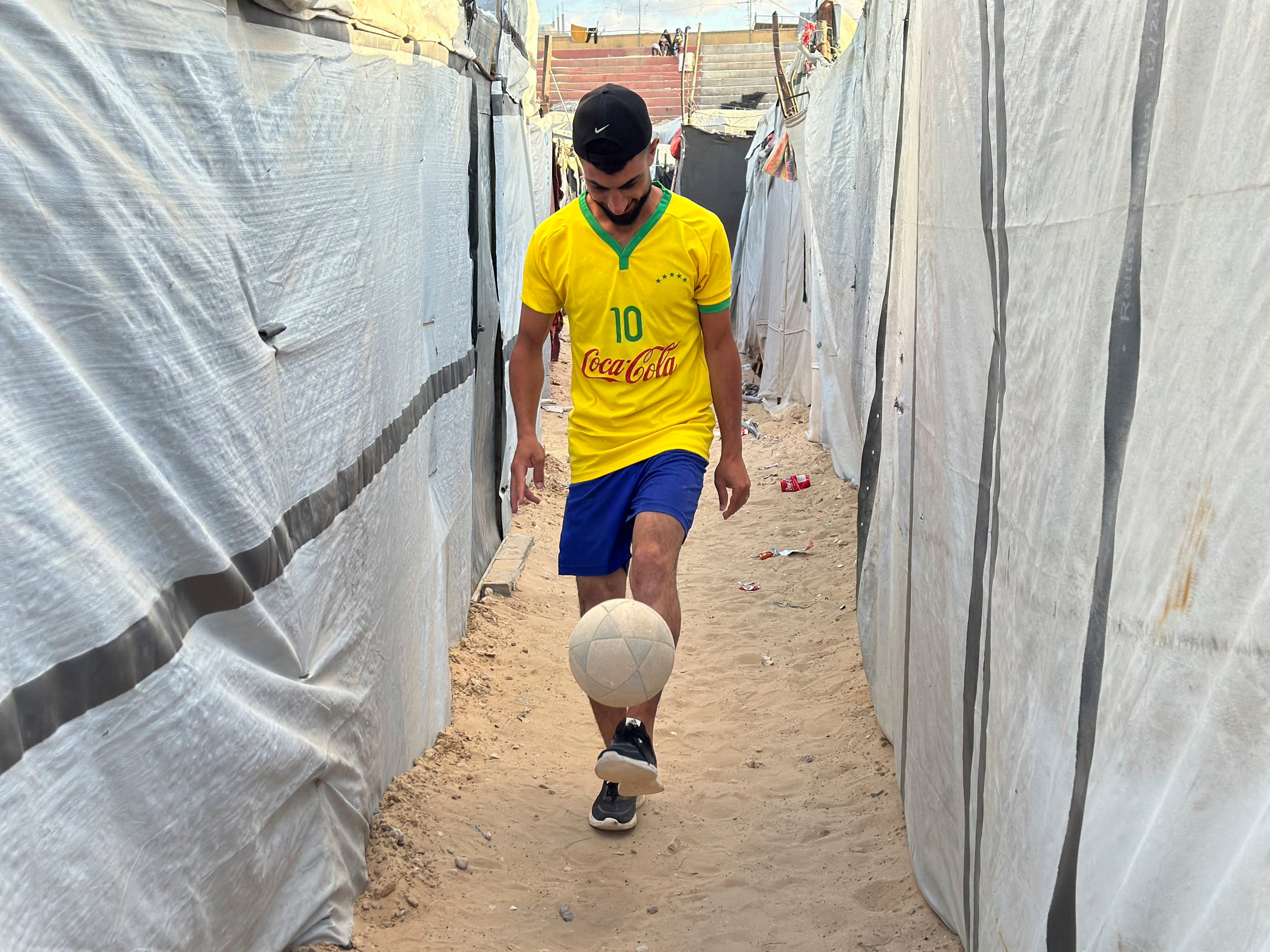A Palestinian football player moves with a ball between tents sheltering displaced Palestinians at the war-damaged Khan Yunis Services Club in Khan Yunis, Gaza Strip, on June 22, 2026 The Khan Yunis Services Club, once one of southern Gaza's main sports clubs, has been turned into a shelter for displaced families after sustaining major damage during the war.