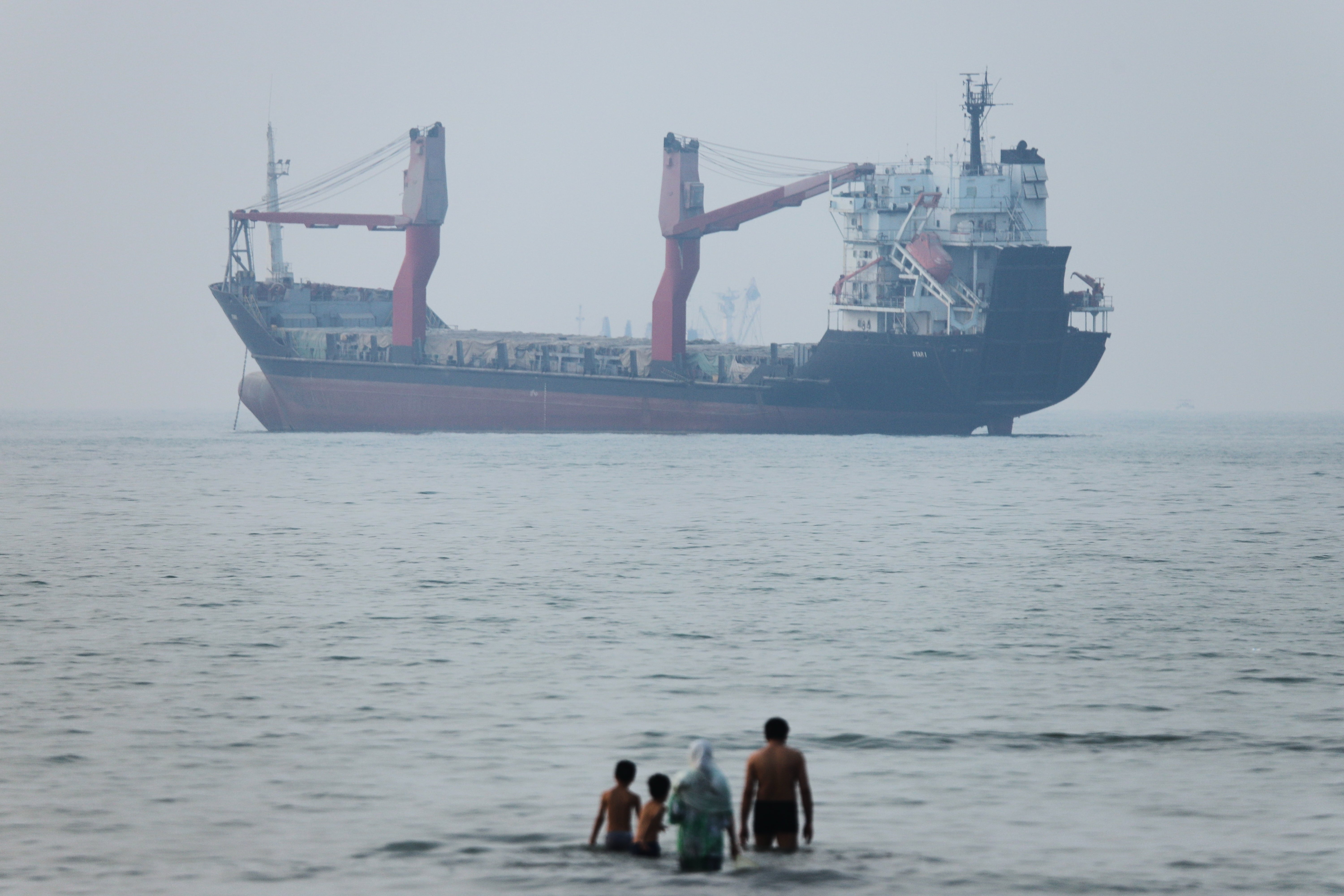 A group of people stand in shallow water as a cargo ship appears anchored in the Strait of Hormuz off Bandar Abbas