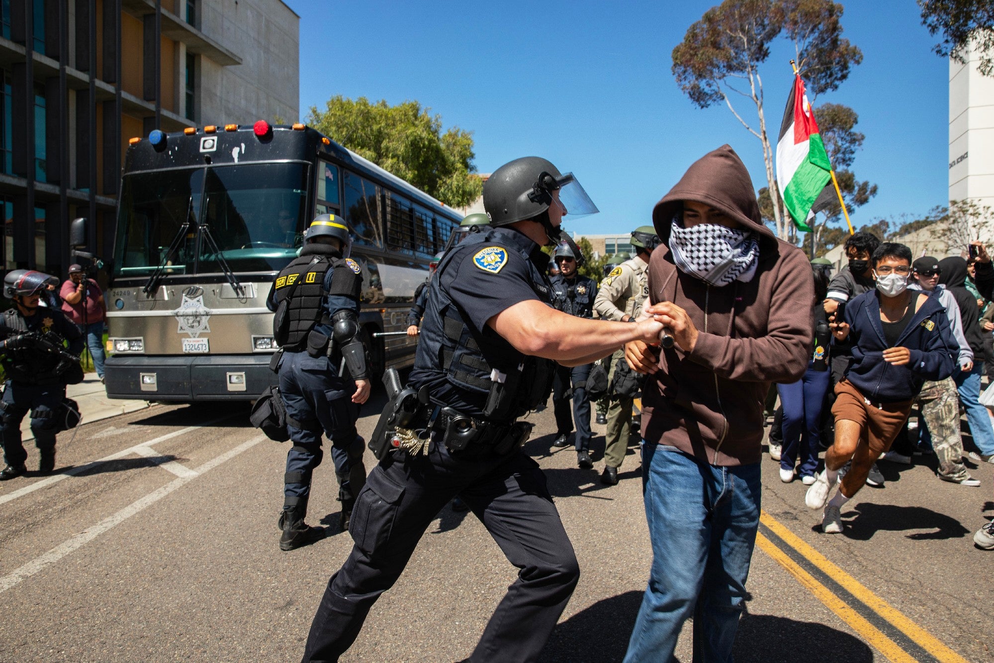 A University of California campus police officer pushes a pro-Palestinian protester away from a moving San Diego Sheriff's bus carrying arrested protesters at UC San Diego, May 6, 2024
