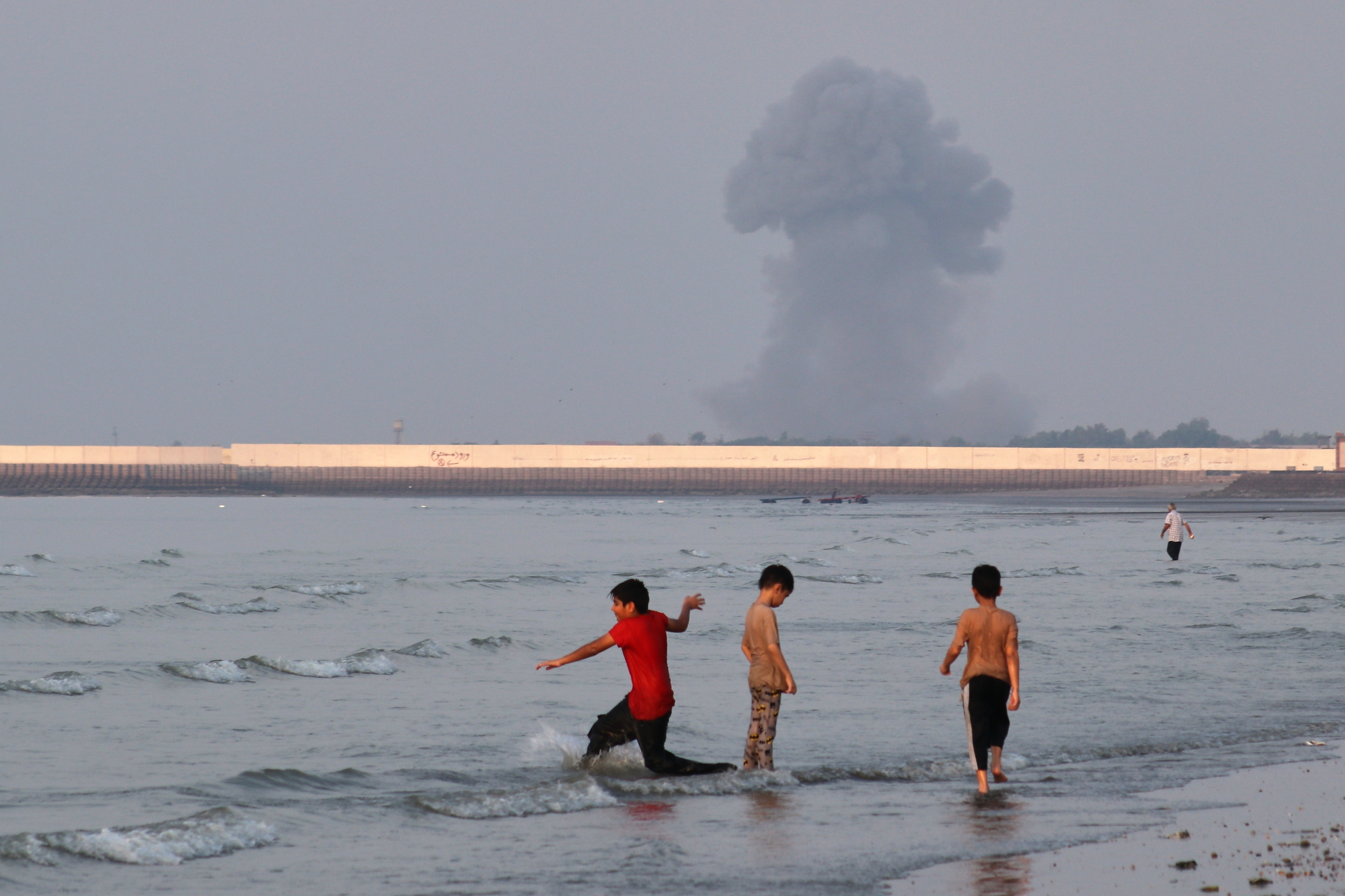 Three boys play in the shallow waters of the Strait of Hormuz, as a plume of smoke rises from an explosion in the background, off Bandar Abbas, Iran