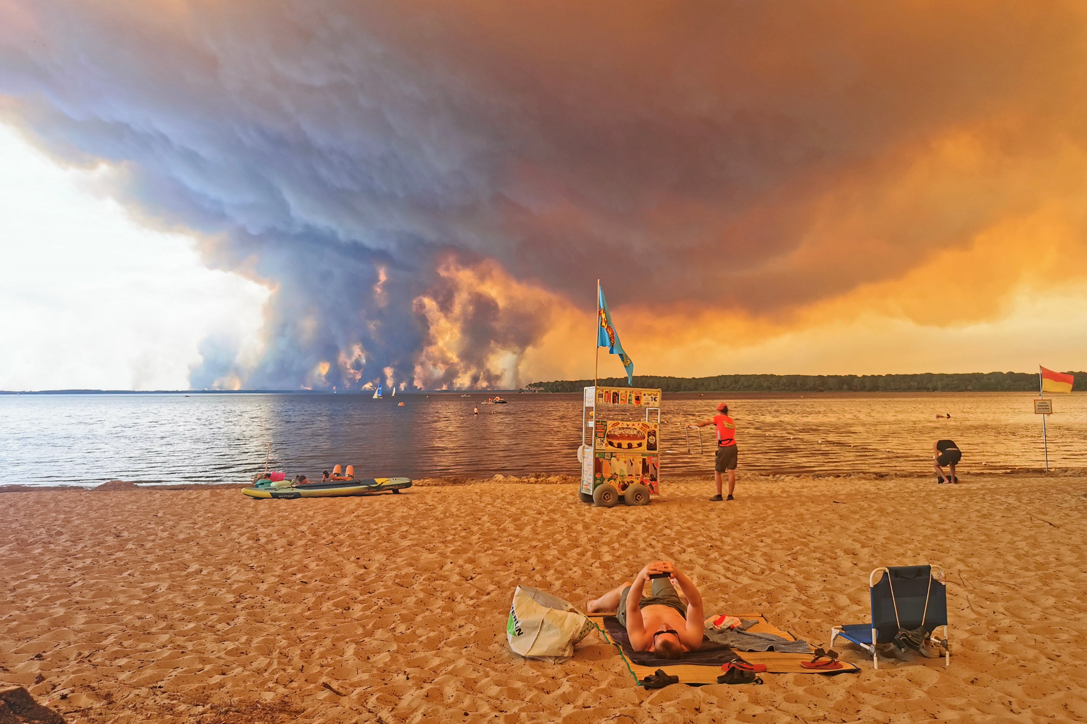 A beach goer looks at his smartphone as a vendor waits with his cart on the beach of Moutchic on the shore of Lake Lacanau (Etang de Lacanau) in Lacanau , southwestern France on July 24, 2026, as clouds of smoke rise into the sky from the Gironde wildfire.