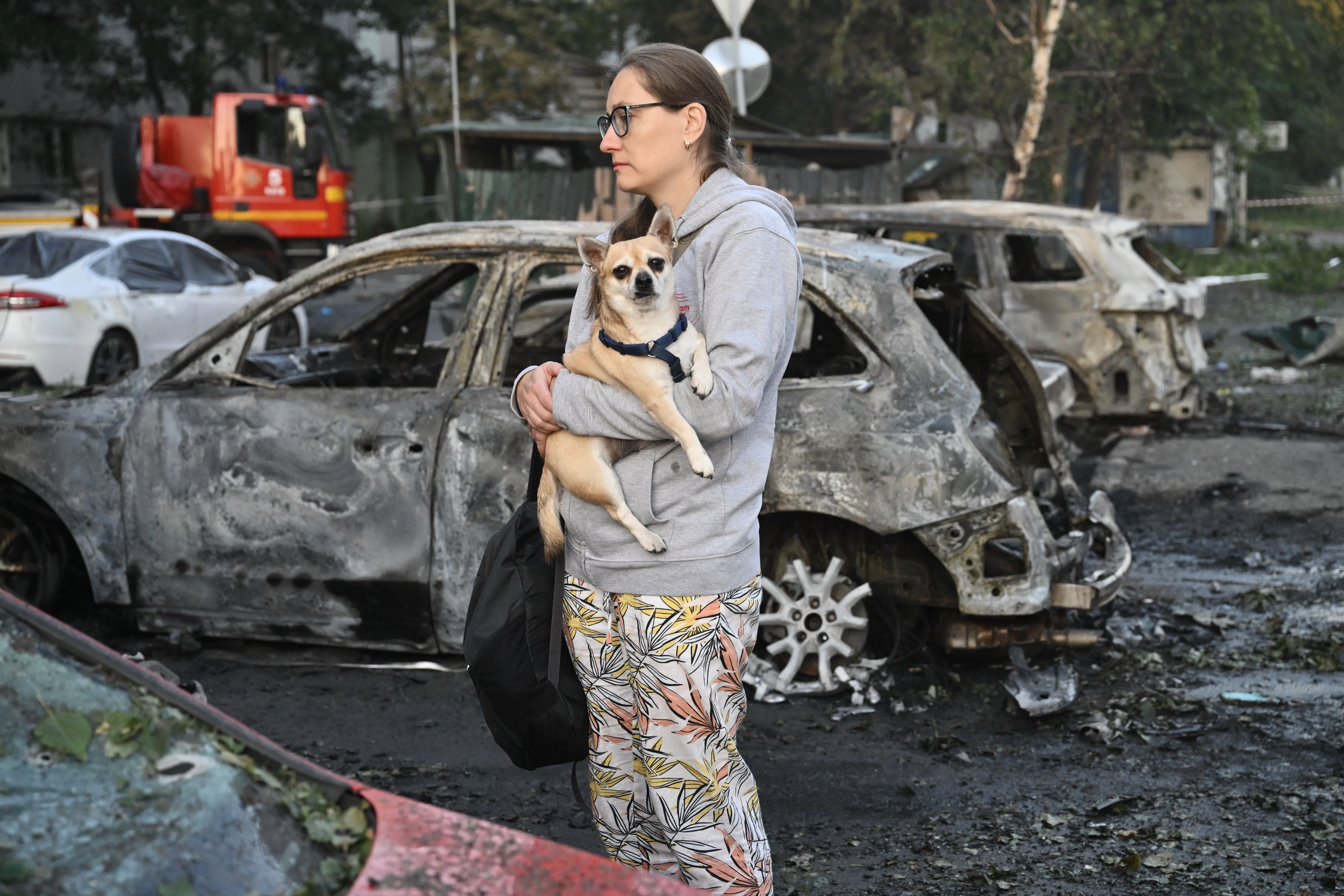 A local resident holds her dog as she examines a broken car parked outside a multi-storey residential building