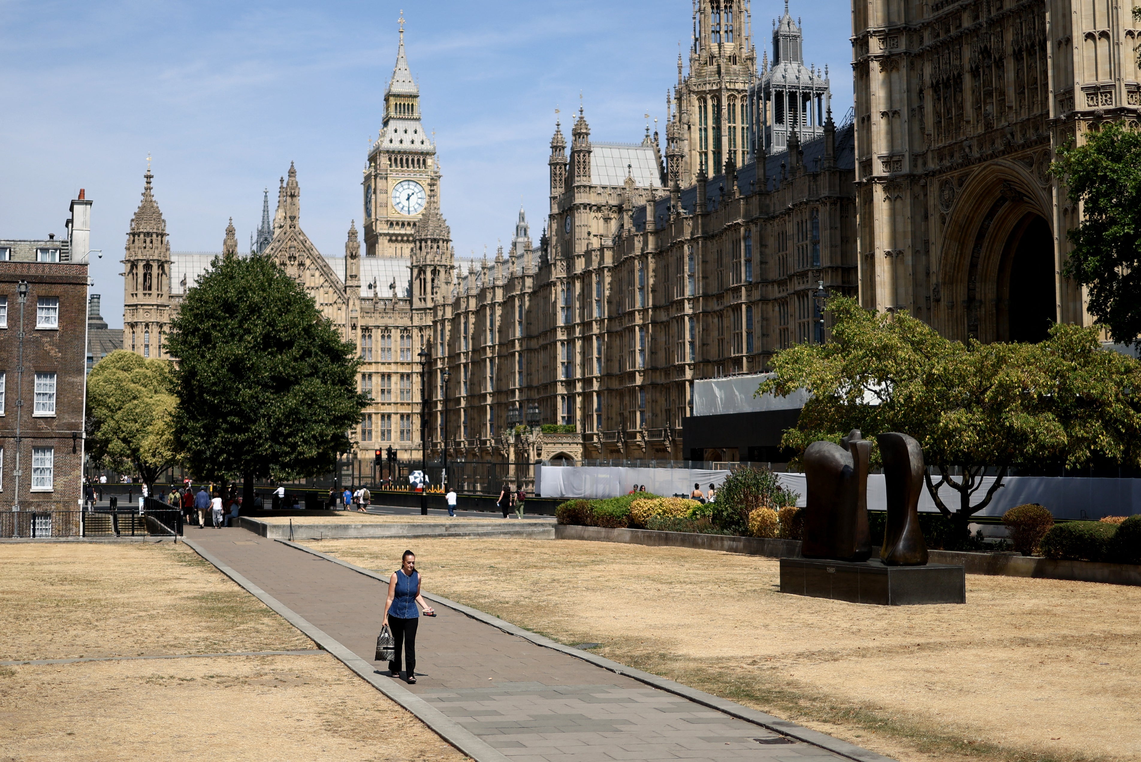 A person walks past sun-scorched grass, backdropped by the Houses of Parliament, after a month without rainfall