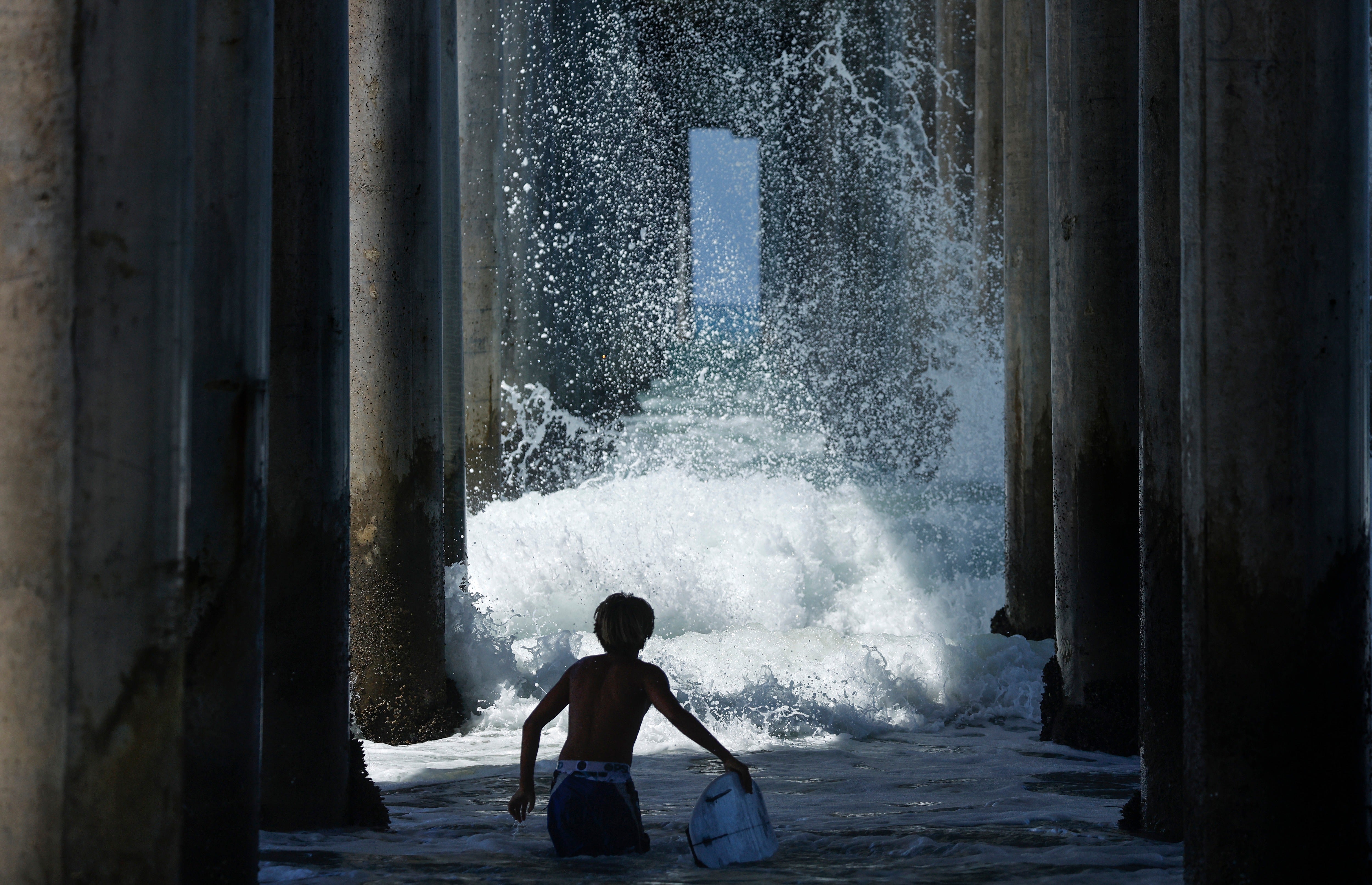 A surfer watches a wave break beneath a pier last month in Huntington Beach, California. Genevieve brought large swells and flooding to Southern California