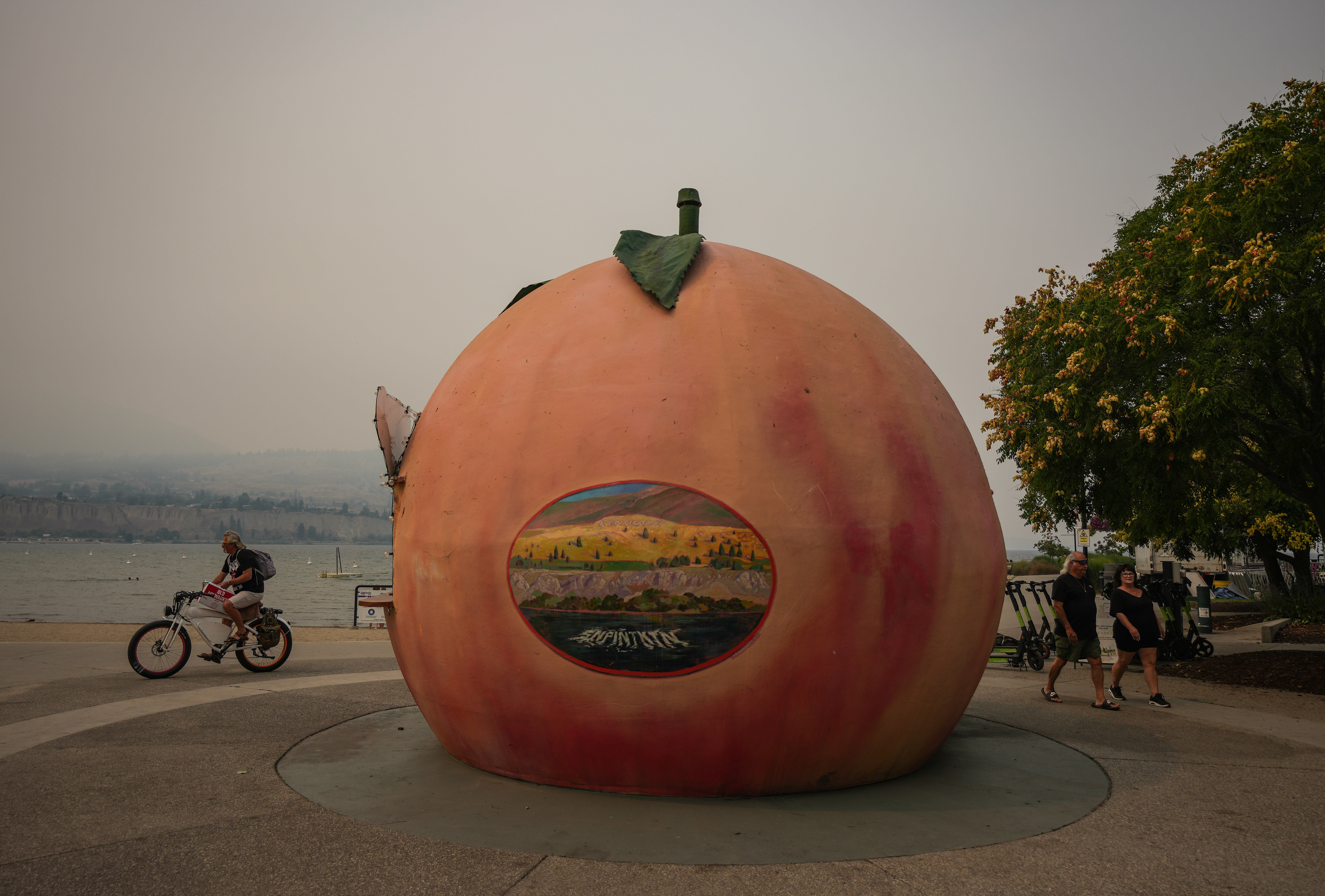 People walk and cycle past an ice cream stand in the shape of a peach as smoke from the Bald Range wildfire envelops Okanagan Lake, in Penticton, British Columbia, on Sunday, Aug. 9, 2026