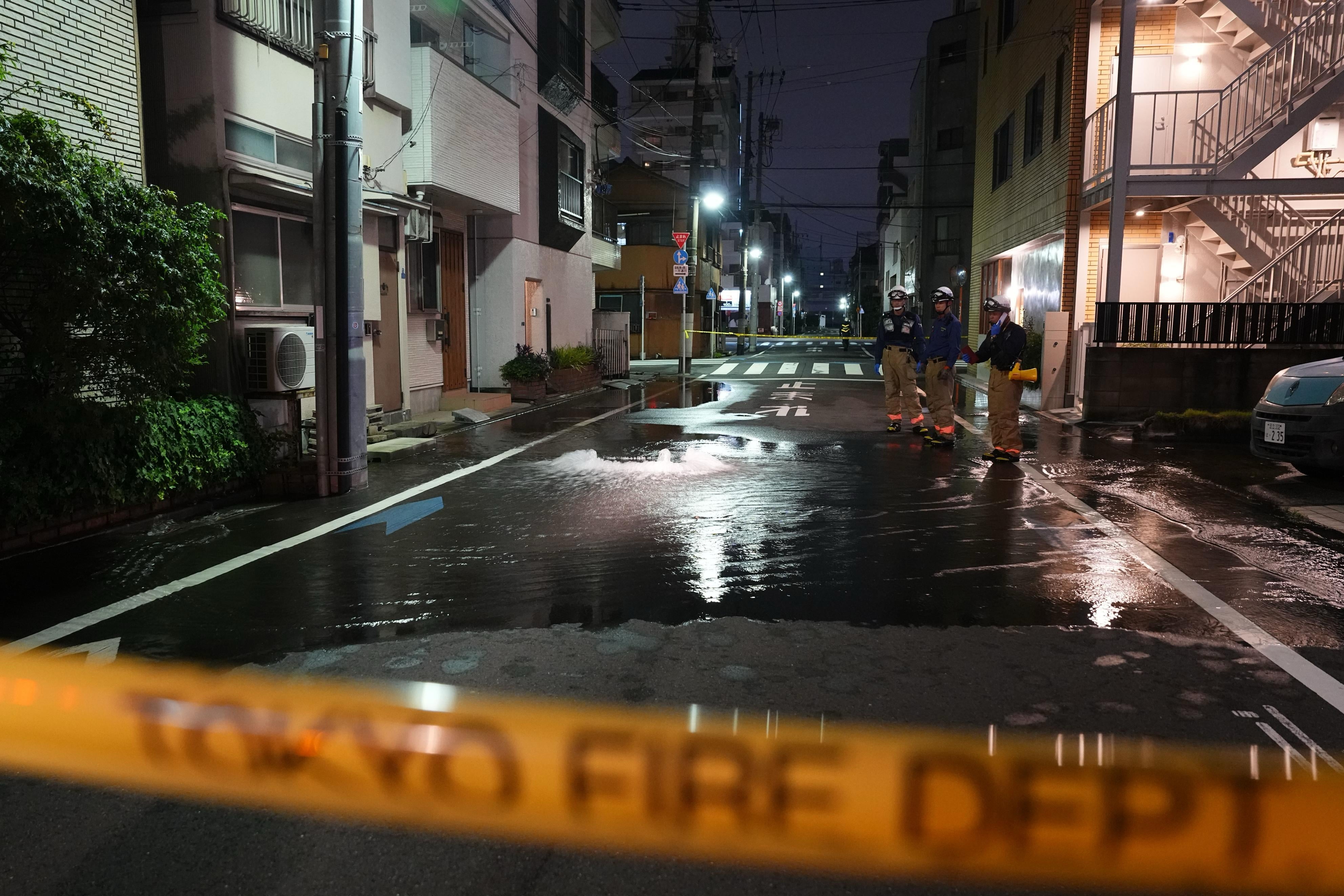 First responders from the Tokyo Fire Department stand on a street partially flooded in the early hours of Sunday, 23 August 2026, following the rupture of a water pipe after an earthquake