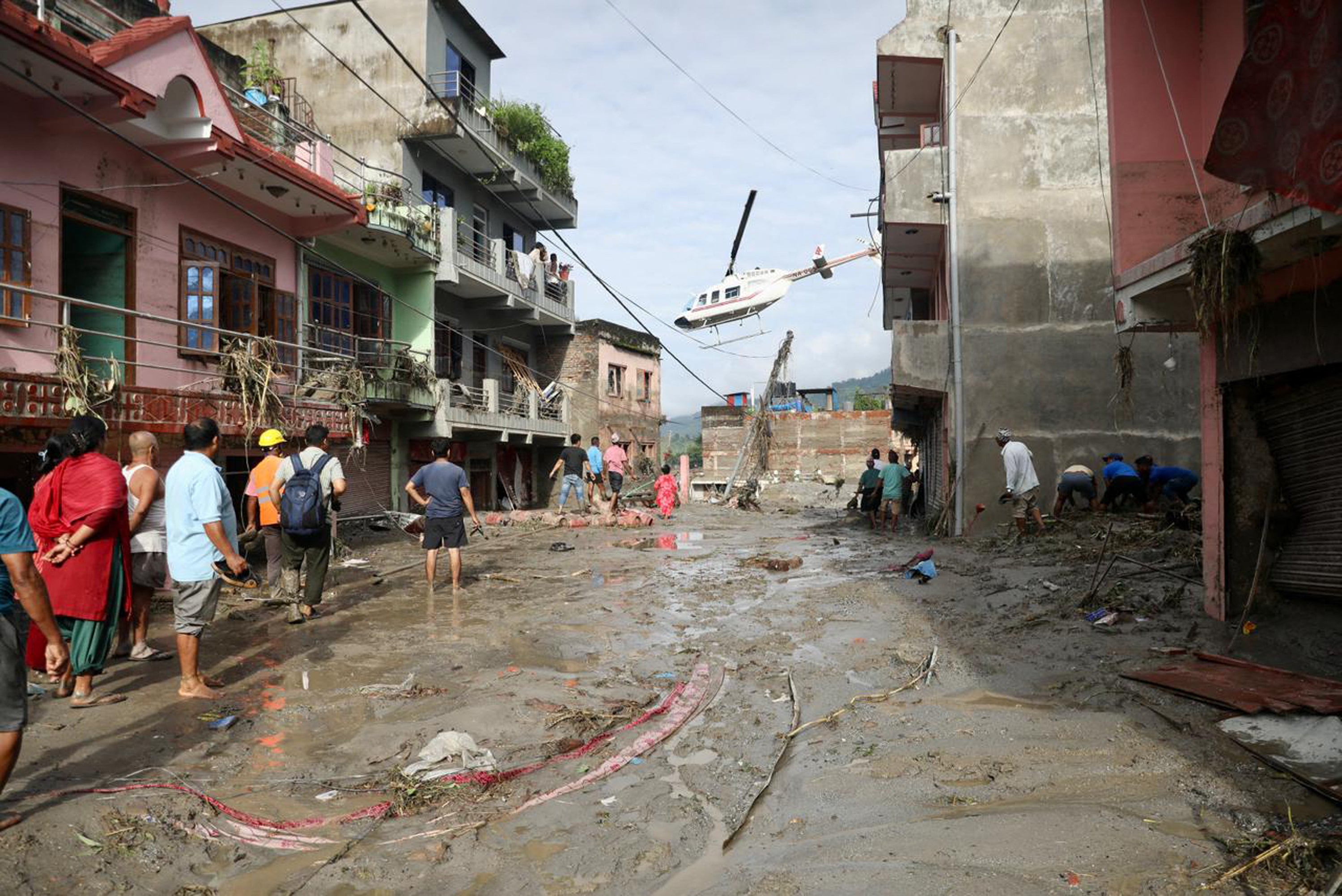 Local residents watch as a helicopter flies above houses lying deluged in sludge after flash floods at Devighat, Bidur Municipality in Nepal's Nuwakot district on August 27, 2026
