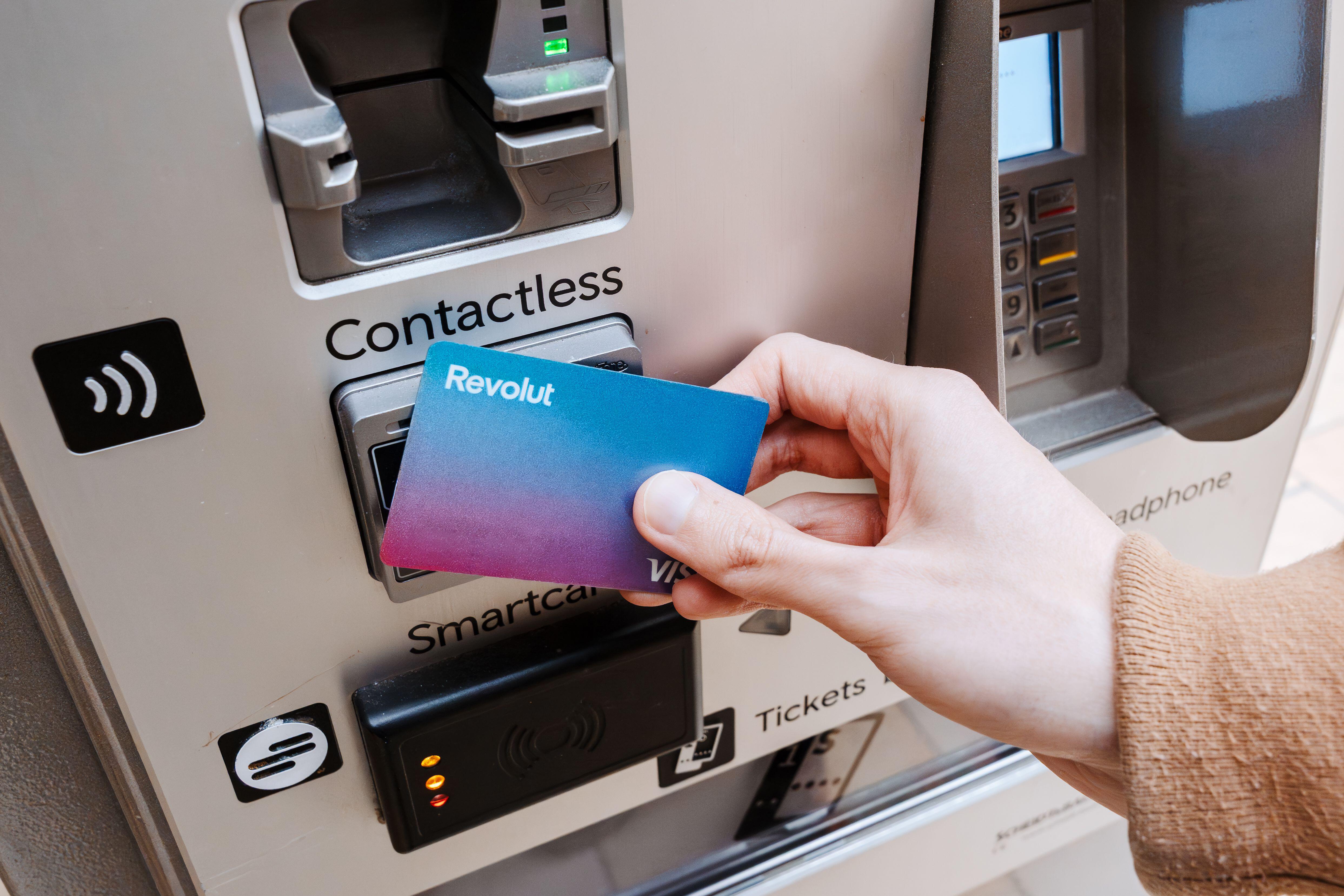 3E7987W London, United Kingdom – 14 March, 2025: Hand holding Revolut Visa bank card tapping contactless payment terminal on automated ticket vending machine,