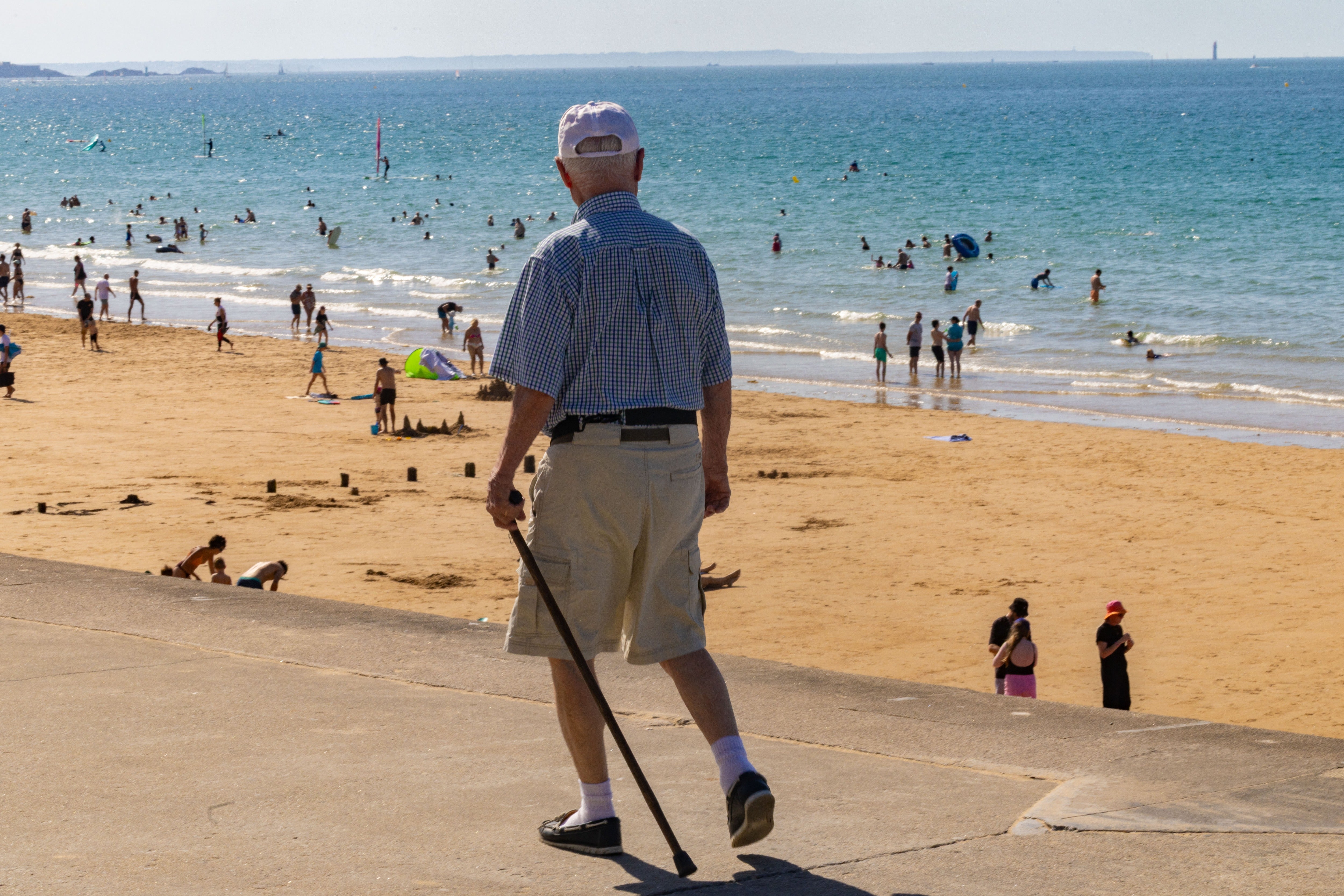 An elderly man walks with a cane, his gaze directed toward the sand